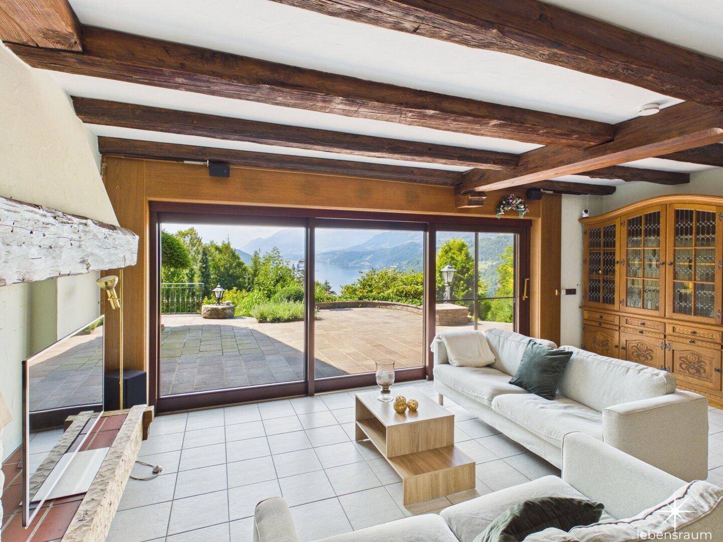 Helles Wohnzimmer mit Holzbalken, Fliesenboden und Panoramafenstern mit atemberaubendem Seeblick.