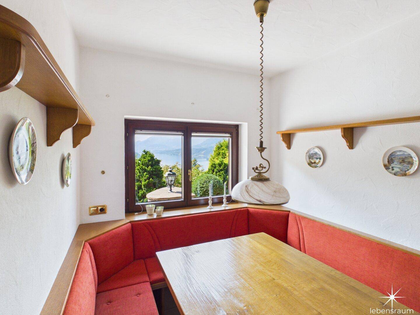 Gemütliche Essecke mit roter Eckbank, Holztisch und malerischem Seeblick durch das Fenster.