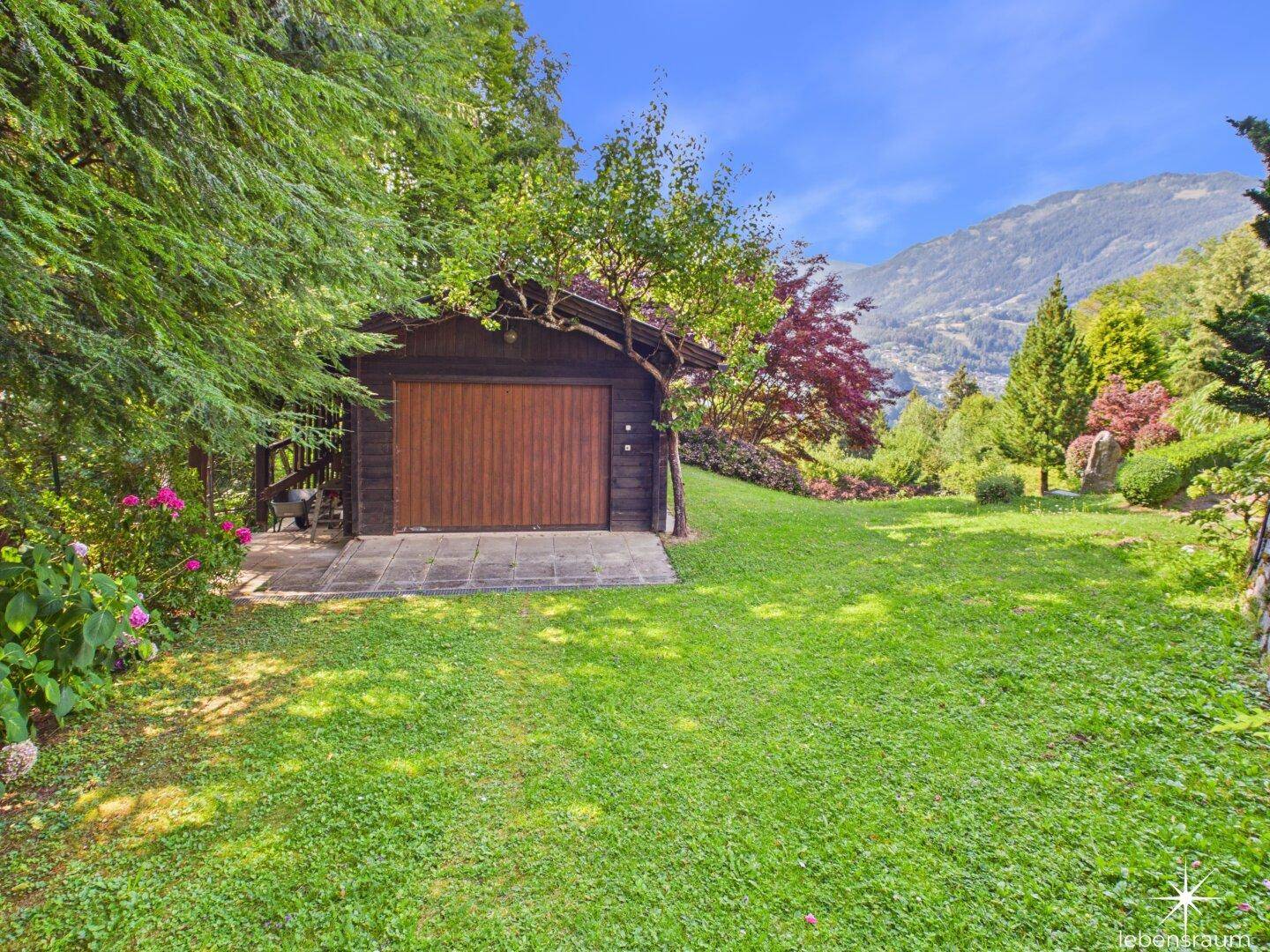Holzgarage oder Schuppen auf einer grünen Wiese, umgeben von Bäumen und Bergen.