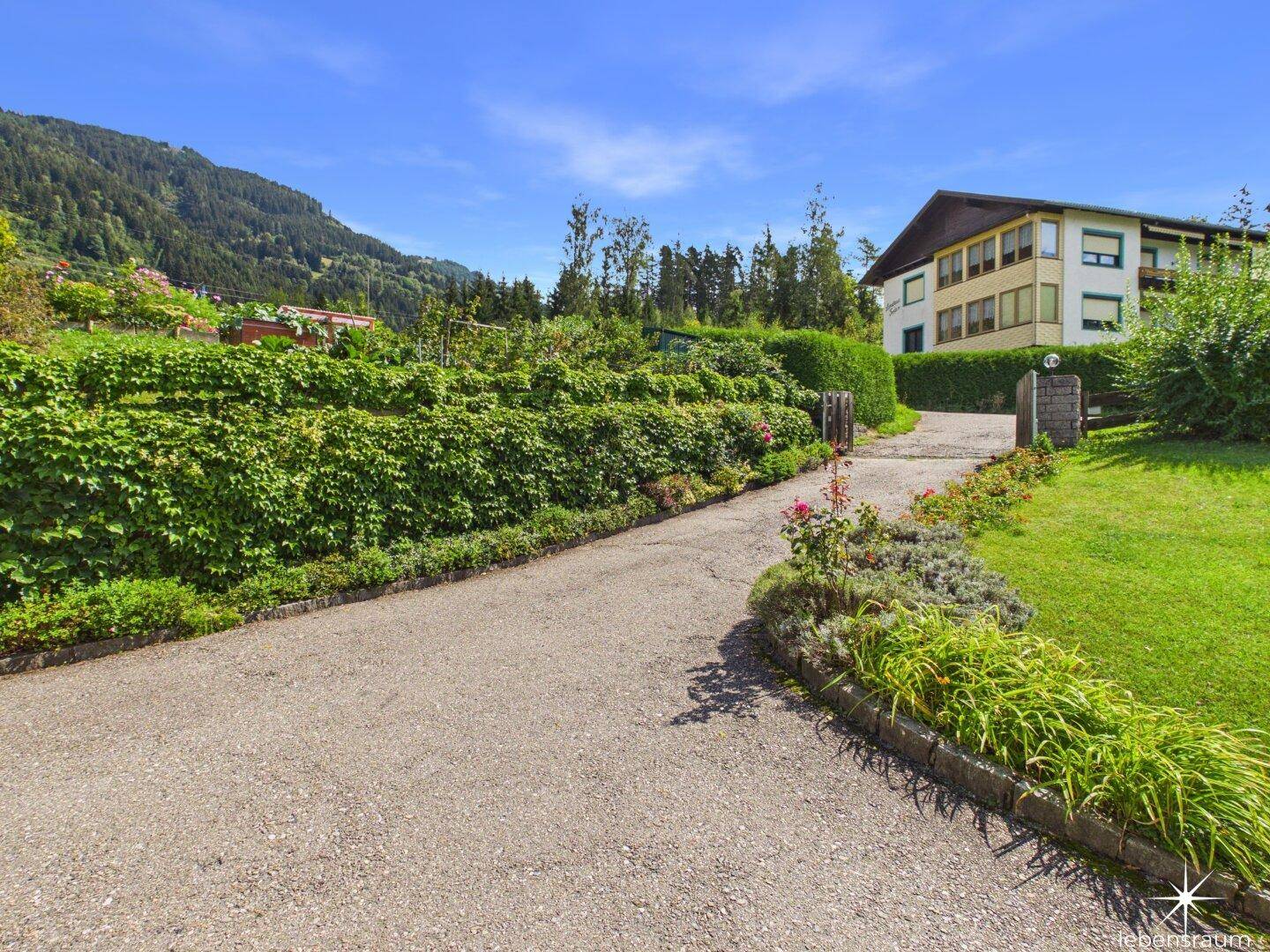 Zufahrt zum Grundstück, gesäumt von Hecken und Blumenbeeten, führt zum Haus.