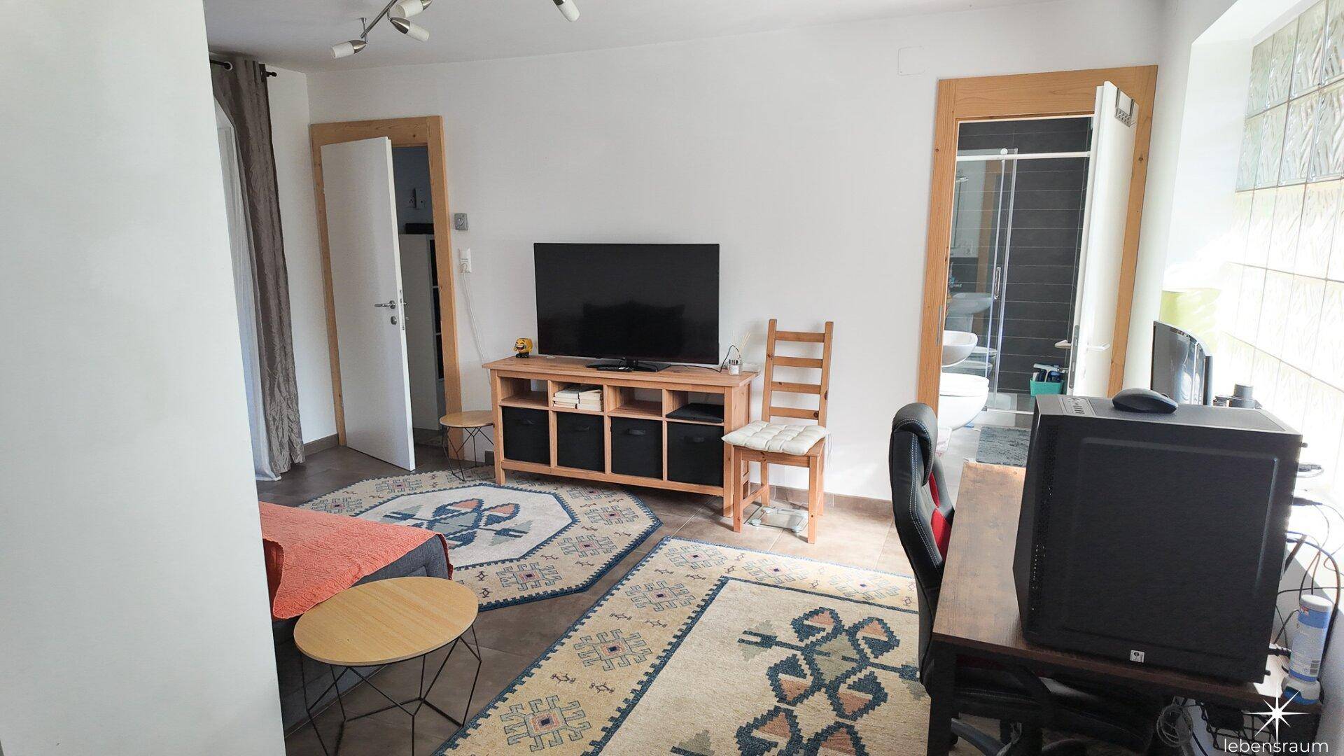 Geräumiges Wohnzimmer mit Arbeitsbereich, Fernseher und Blick auf ein angrenzendes Badezimmer.