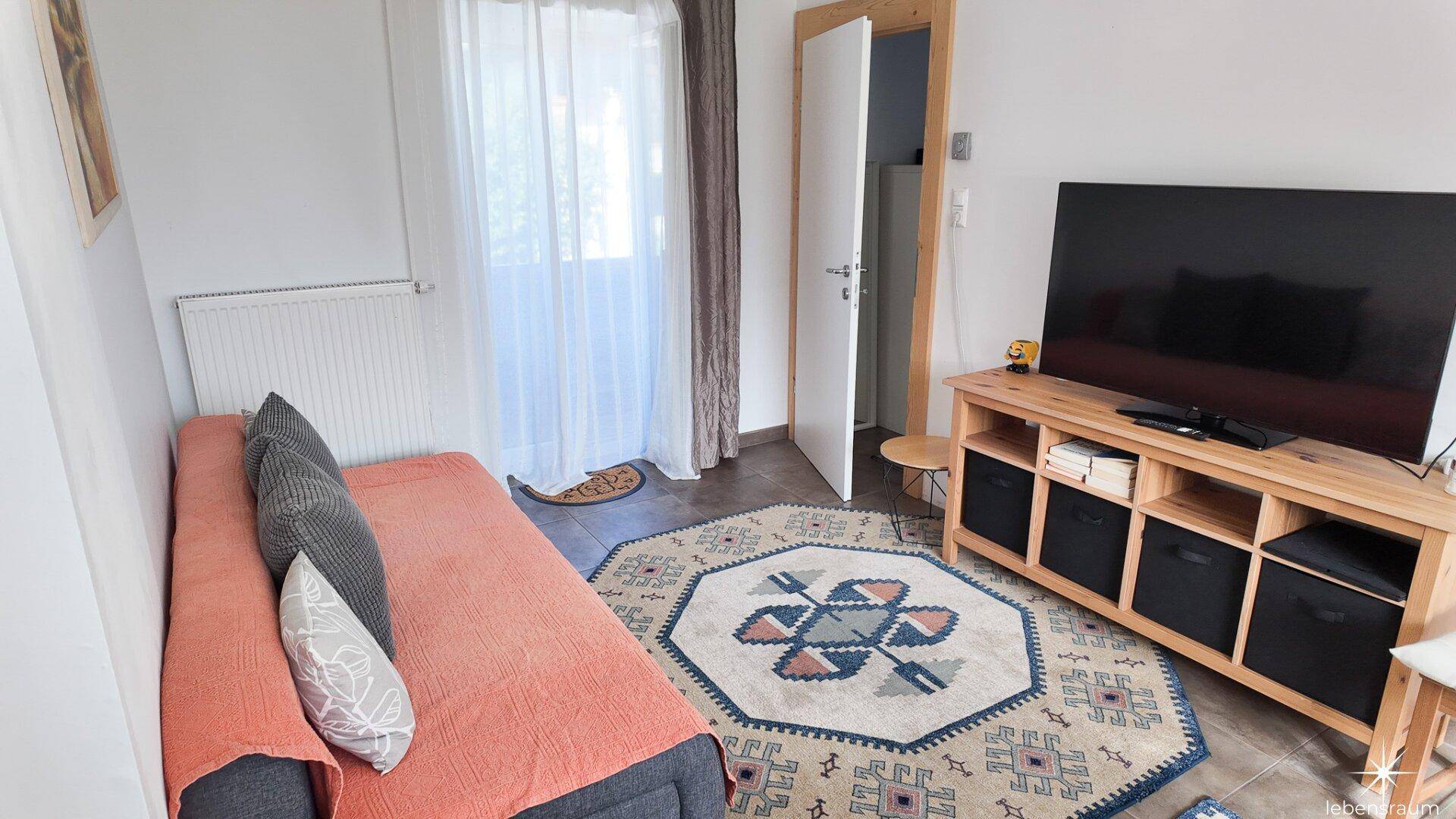 Helles Wohnzimmer mit gemütlichem Sofa, Fernseher und Zugang zu einem Balkon oder einer Terrasse.