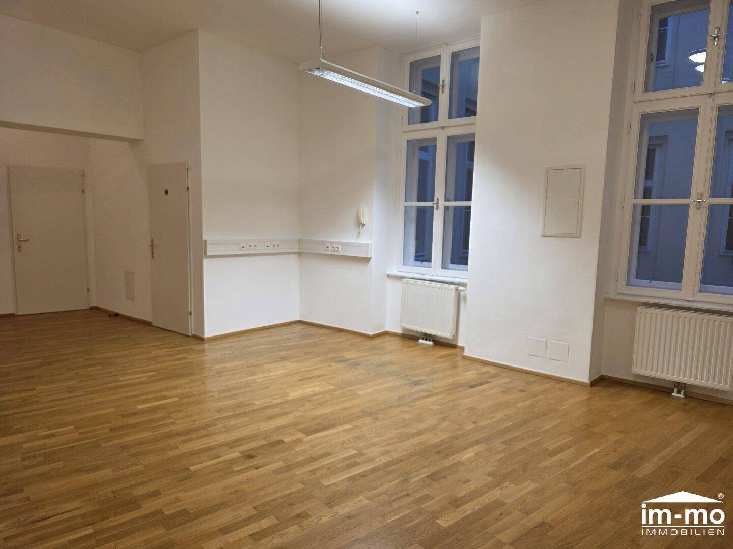 Geräumiger Büroraum mit Holzboden, großen Fenstern und einfacher Beleuchtung.