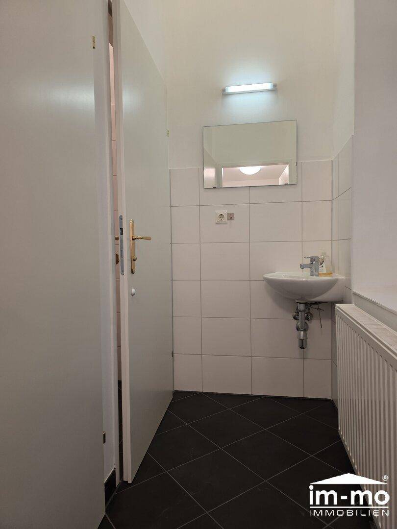 Kleines Badezimmer mit Waschbecken, Spiegel und Heizkörper, gefliest mit dunklem Boden.
