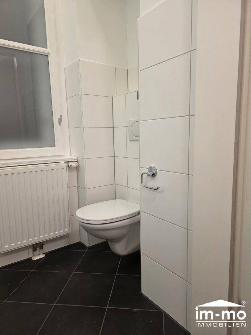 Toilette mit Wand-WC, weißen Fliesen und Heizkörper unter dem Fenster.