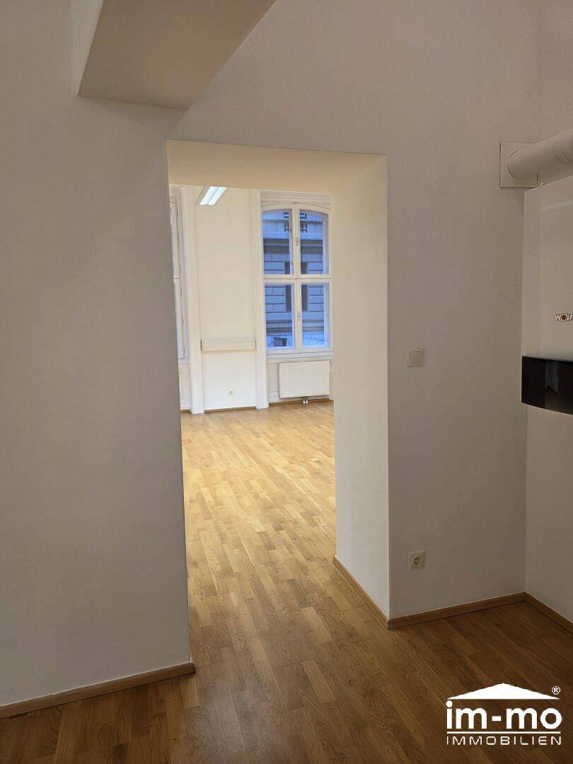 Flur mit Holzboden, der zu einem weiteren hellen Raum mit Fenstern führt.