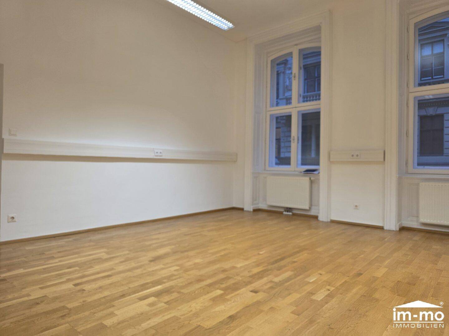 Langer Raum mit Holzboden und zwei großen Fenstern, die den Raum mit Tageslicht füllen.