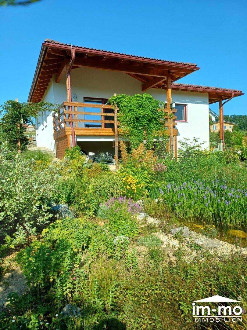 Hausfassade mit großem, gepflegtem Garten und üppiger Bepflanzung unter blauem Himmel.