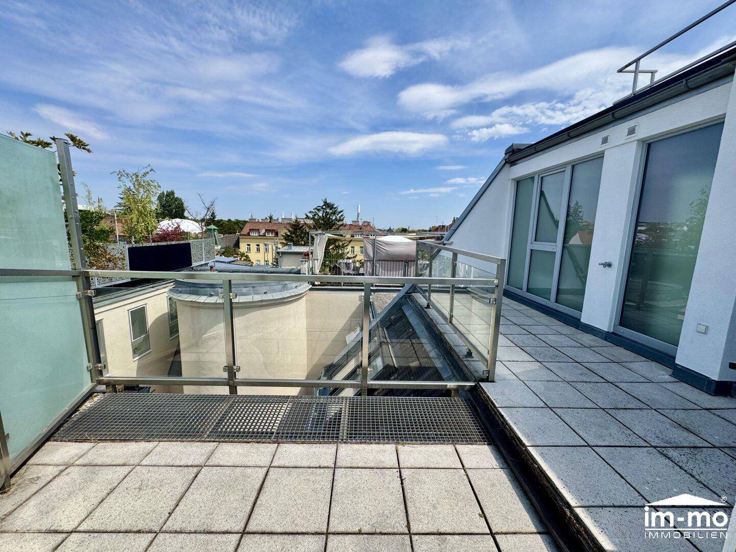 Weitläufige Dachterrasse mit Glasgeländer und Blick auf die umliegenden Gebäude und Grünflächen.