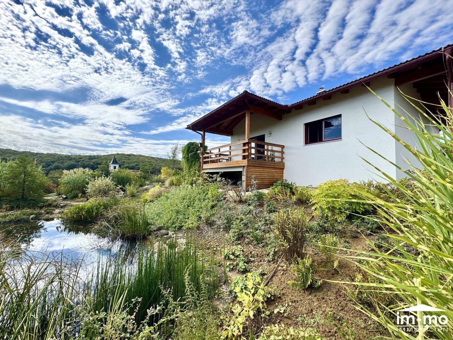 Haus mit Holzbalcon, umgeben von einem naturnahen Garten mit Teich und Fernblick auf die Landschaft.