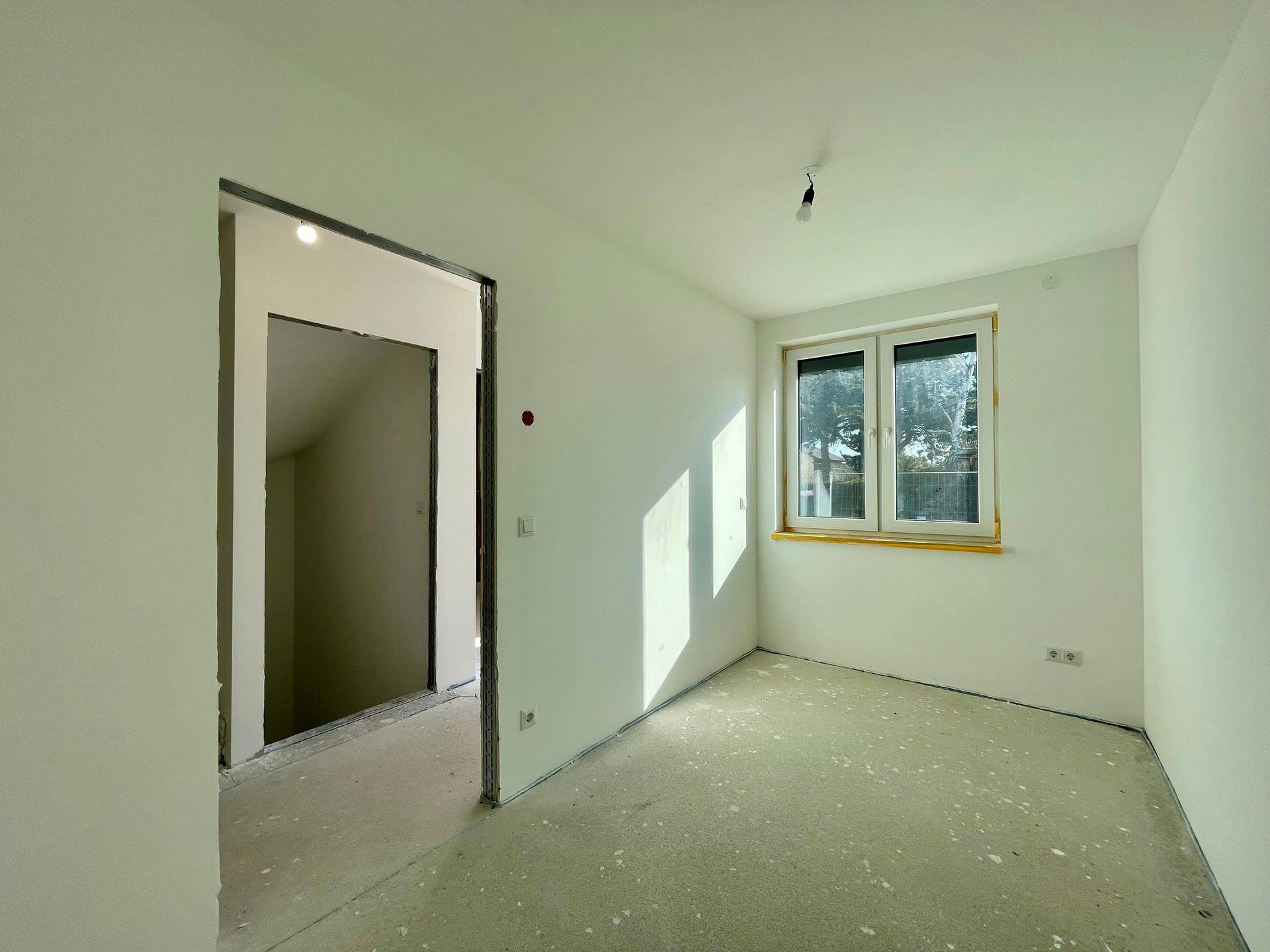 Helles Zimmer im Rohbau mit einem Fenster und Betonboden, bereit für den Ausbau.