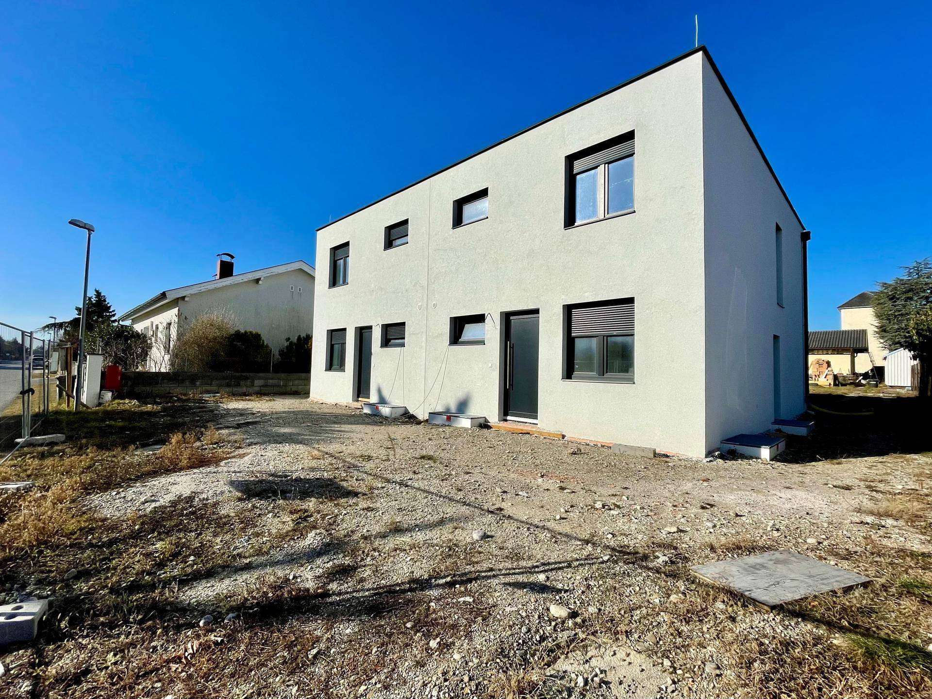 Neubauhaus mit weißer Fassade und dunklen Fenstern, umgeben von unbebautem Grundstück.