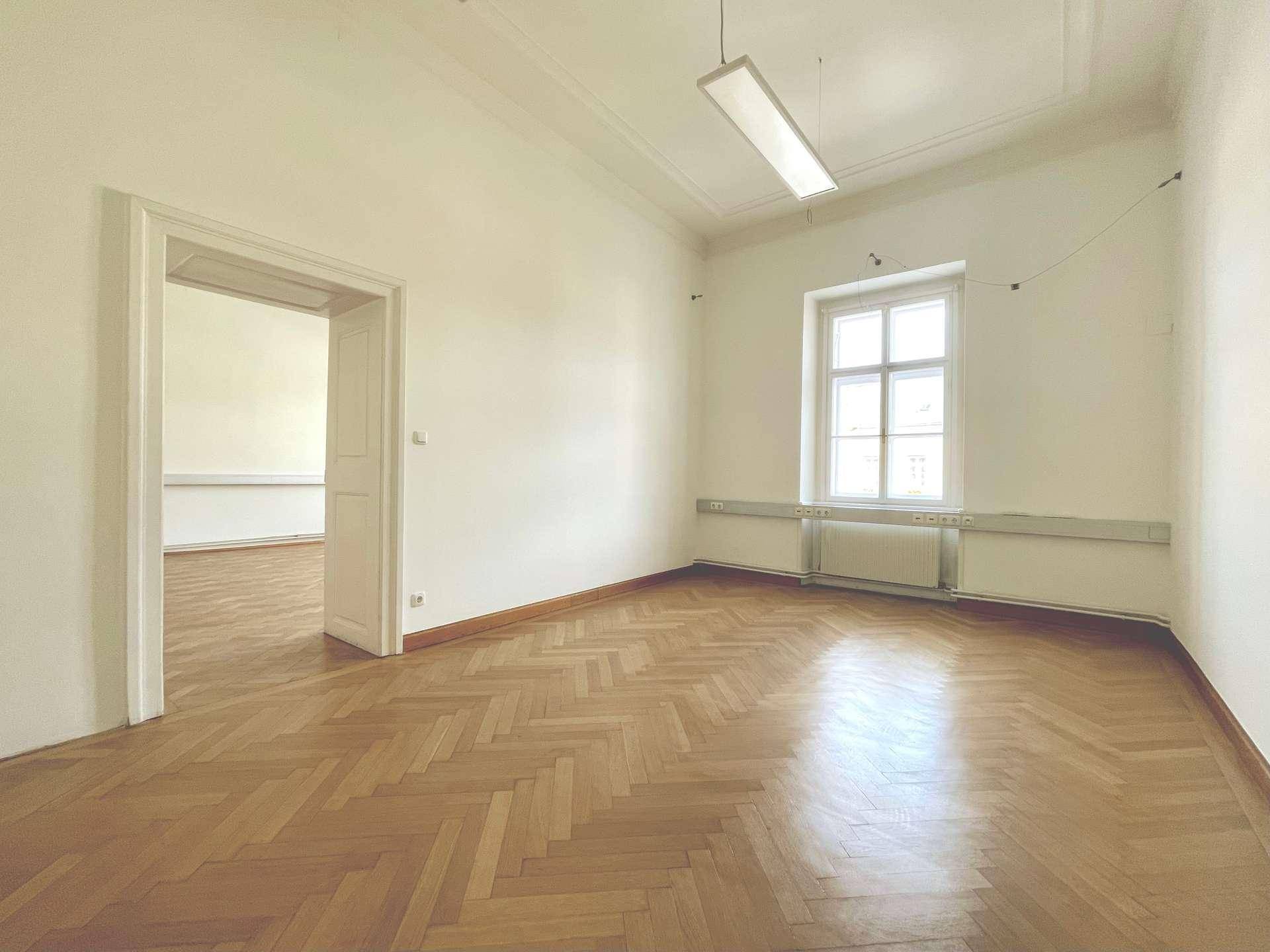 Großer, heller Raum mit Fischgrätparkett und einem Fenster, das viel Tageslicht hereinlässt.