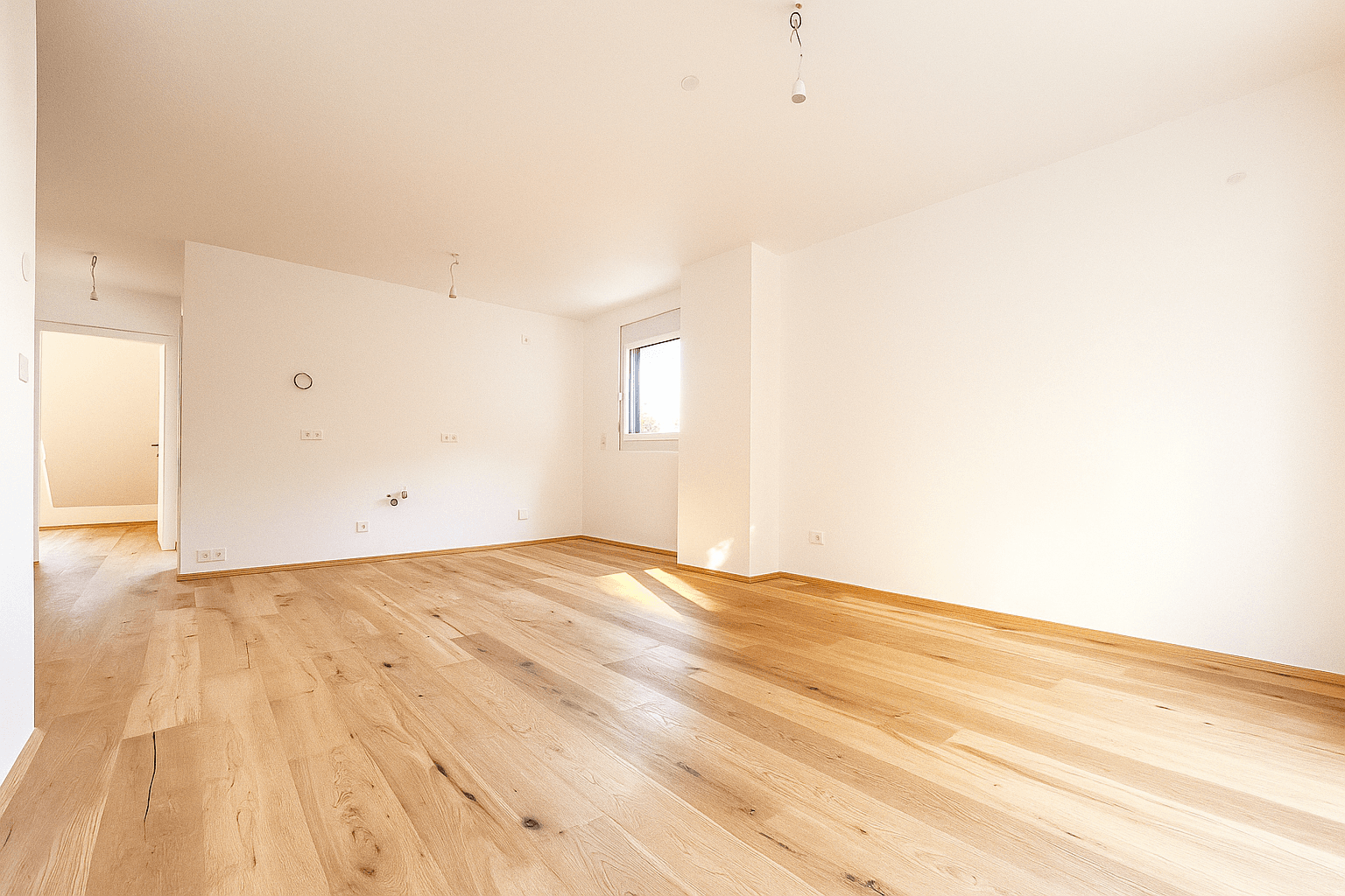 Heller, leerer Wohnraum mit Holzboden und großen Fenstern, die viel Tageslicht hereinlassen.