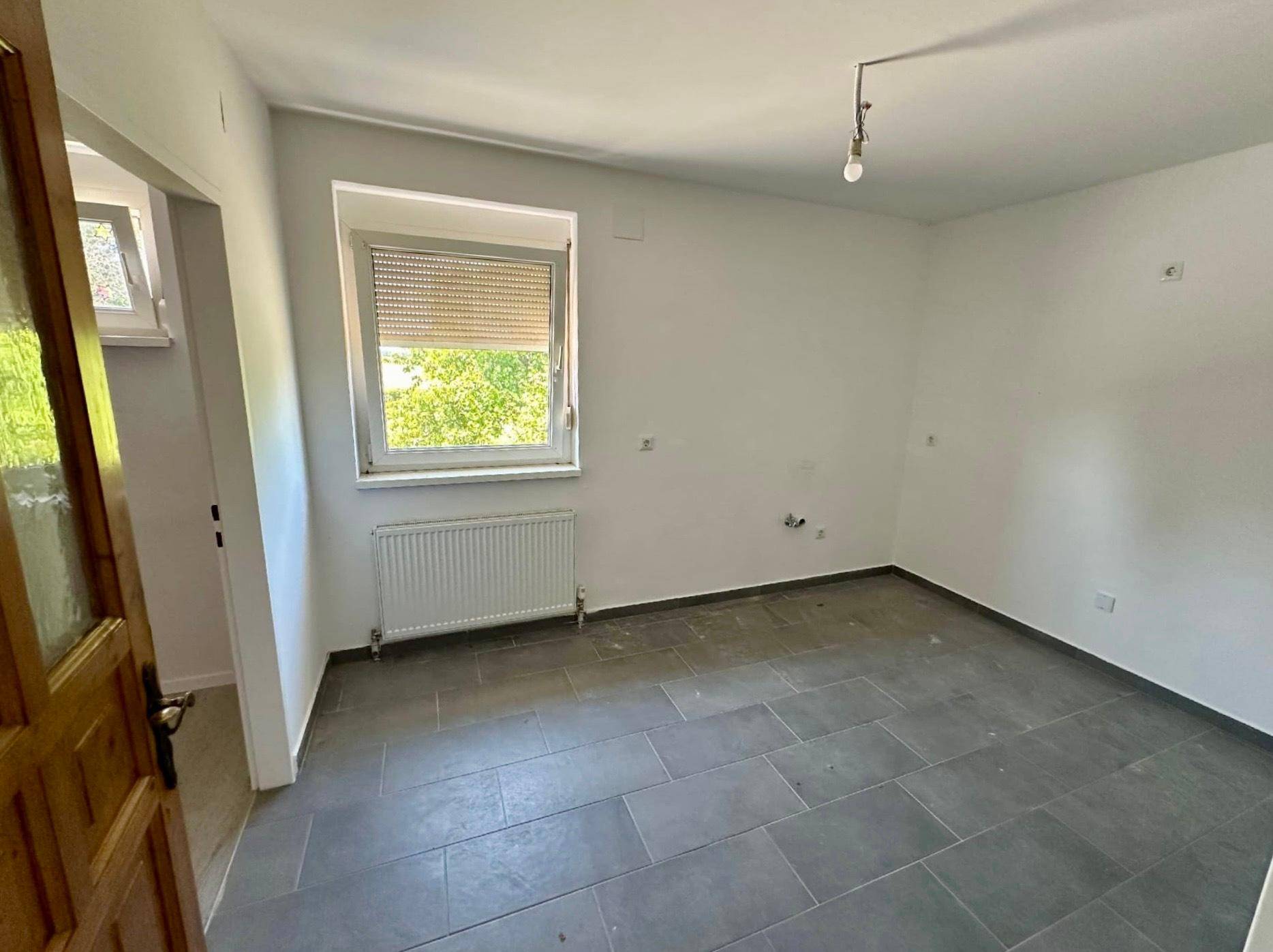 Leeres Zimmer mit grauen Fliesen, Fenster und Heizkörper, bereit zur individuellen Gestaltung und Renovierung.