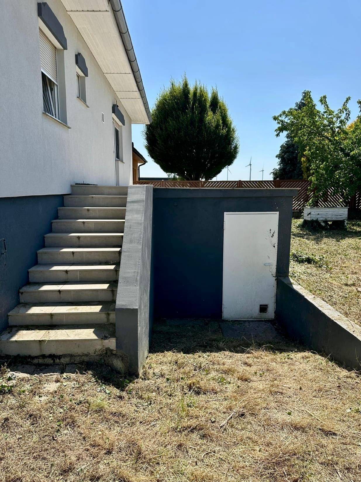 Außentreppe führt zu einem Kellerzugang an der Seite des Hauses, umgeben von trockenem Gras.