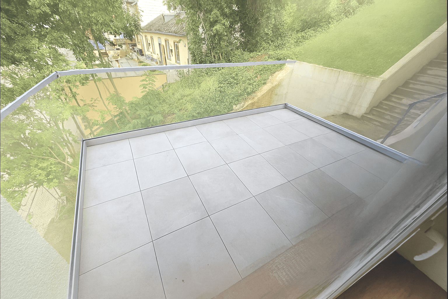 Kleiner Balkon mit Fliesenboden und Glasgeländer, bietet einen Blick auf die grüne Umgebung.