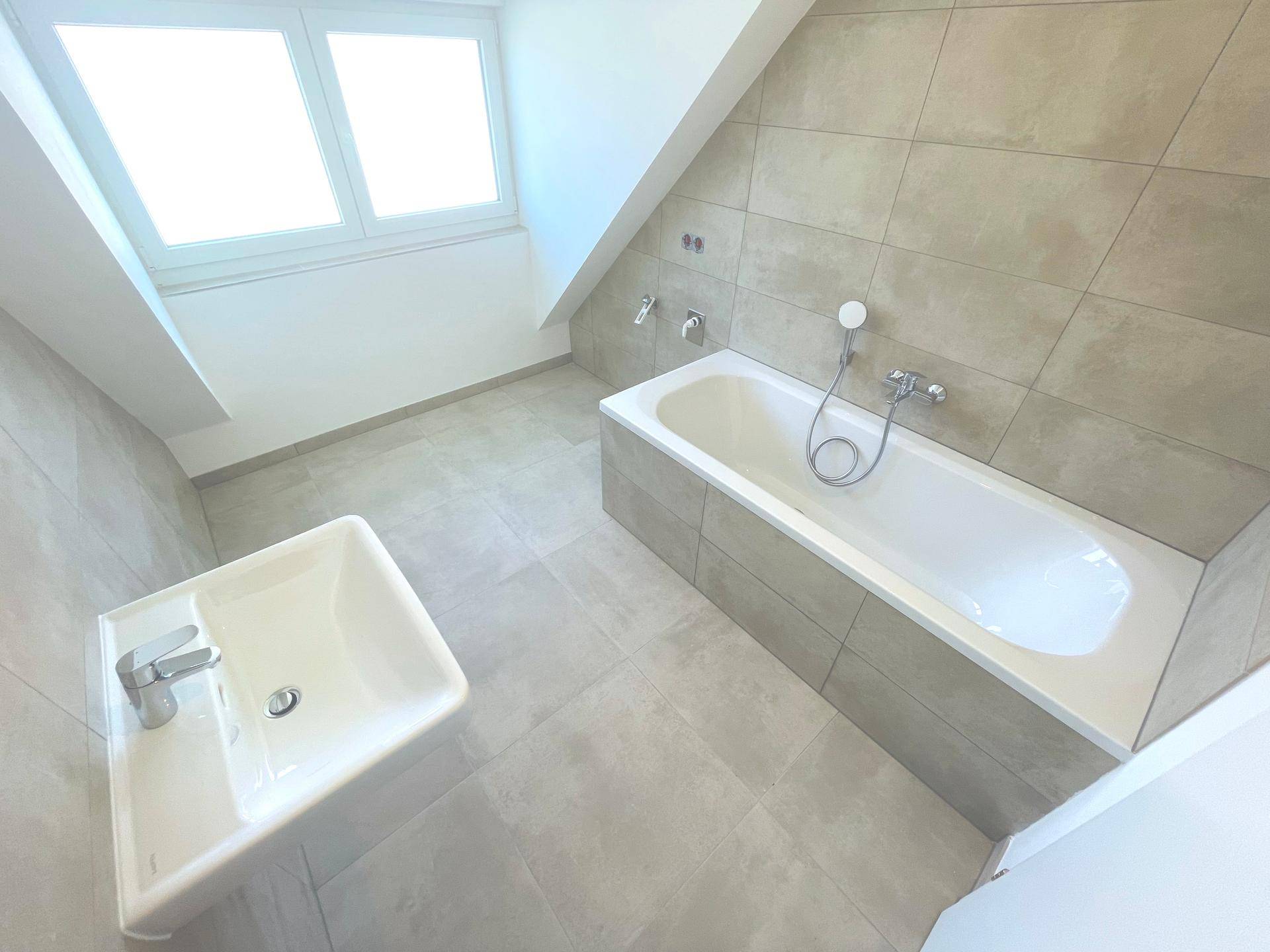 Modernes Badezimmer im Dachgeschoss mit Badewanne, Waschbecken und Fenster, ausgestattet mit hellen Fliesen.