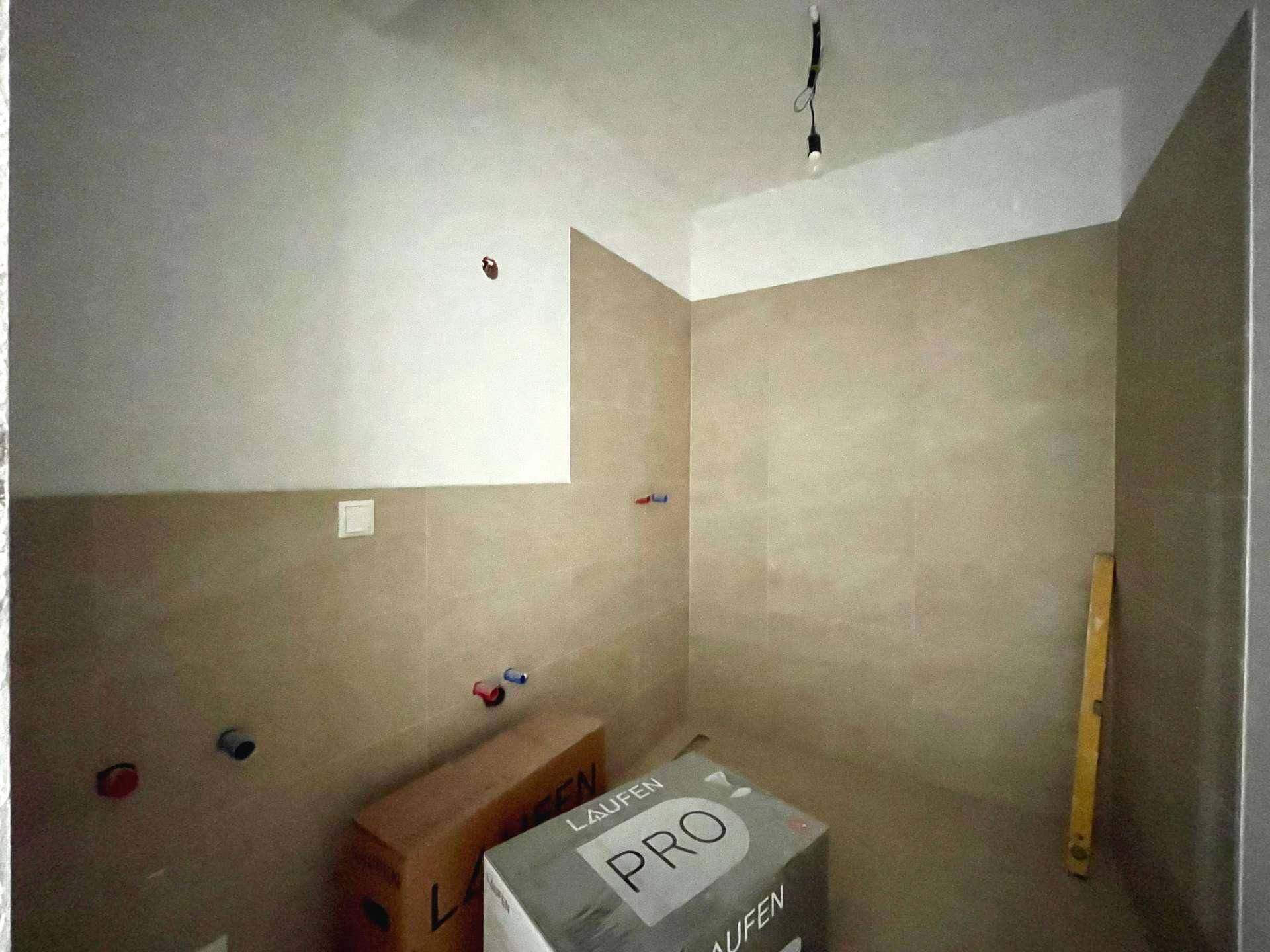 Badezimmer im Rohbau mit beigefarbenen Fliesen und vorbereiteten Anschlüssen für Sanitärobjekte.