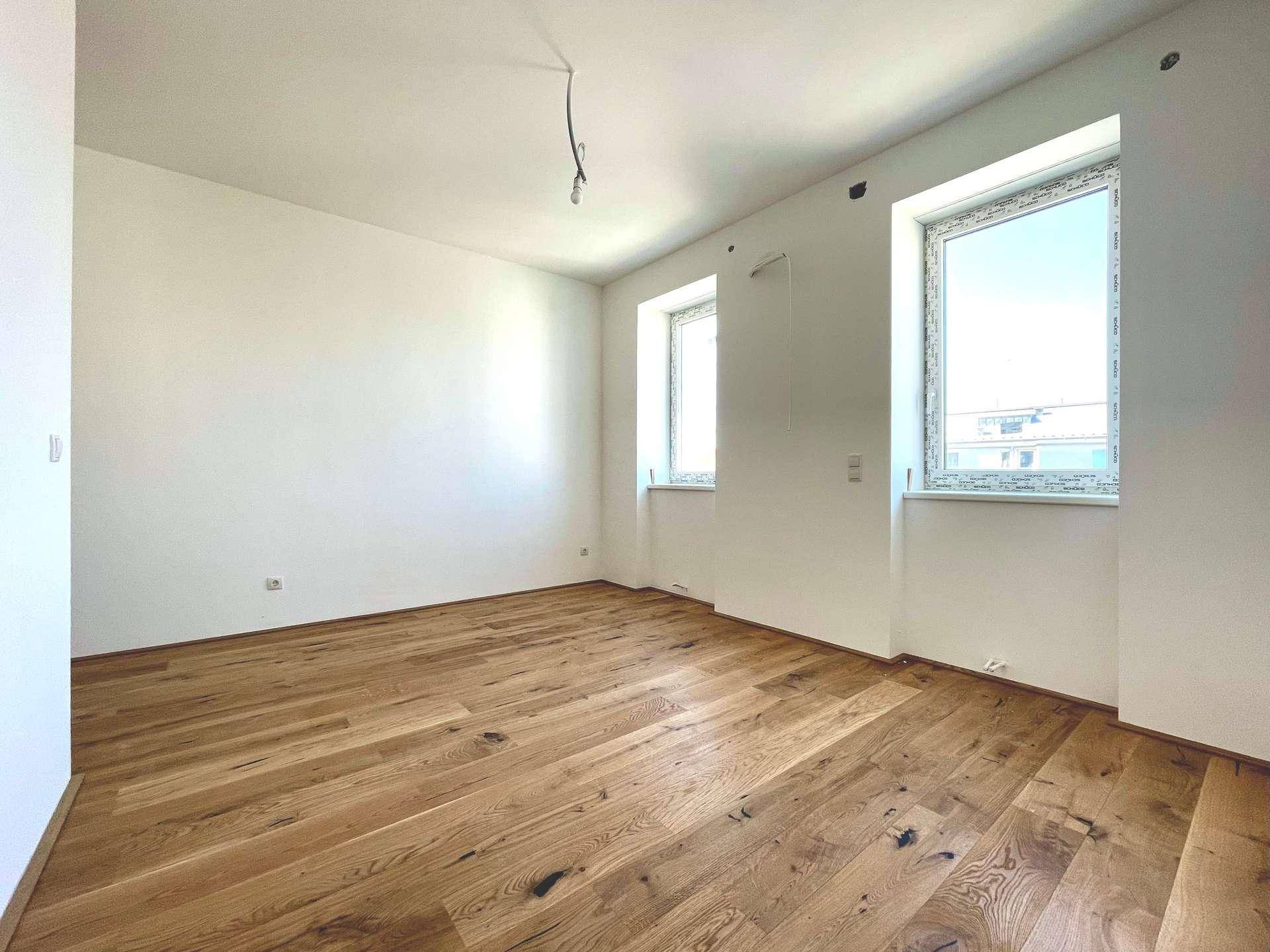 Lichtdurchfluteter Raum mit Holzboden und zwei Fenstern, ideal als Schlafzimmer oder Arbeitszimmer.