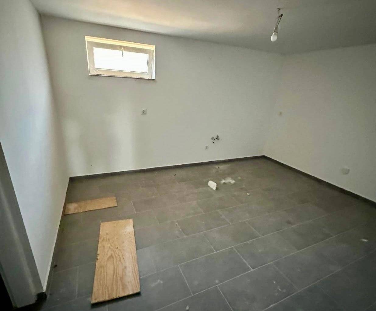 Leeres Zimmer mit grauen Fliesen und kleinem Fenster, ideal für ein Gästezimmer oder Büro nach Renovierung.