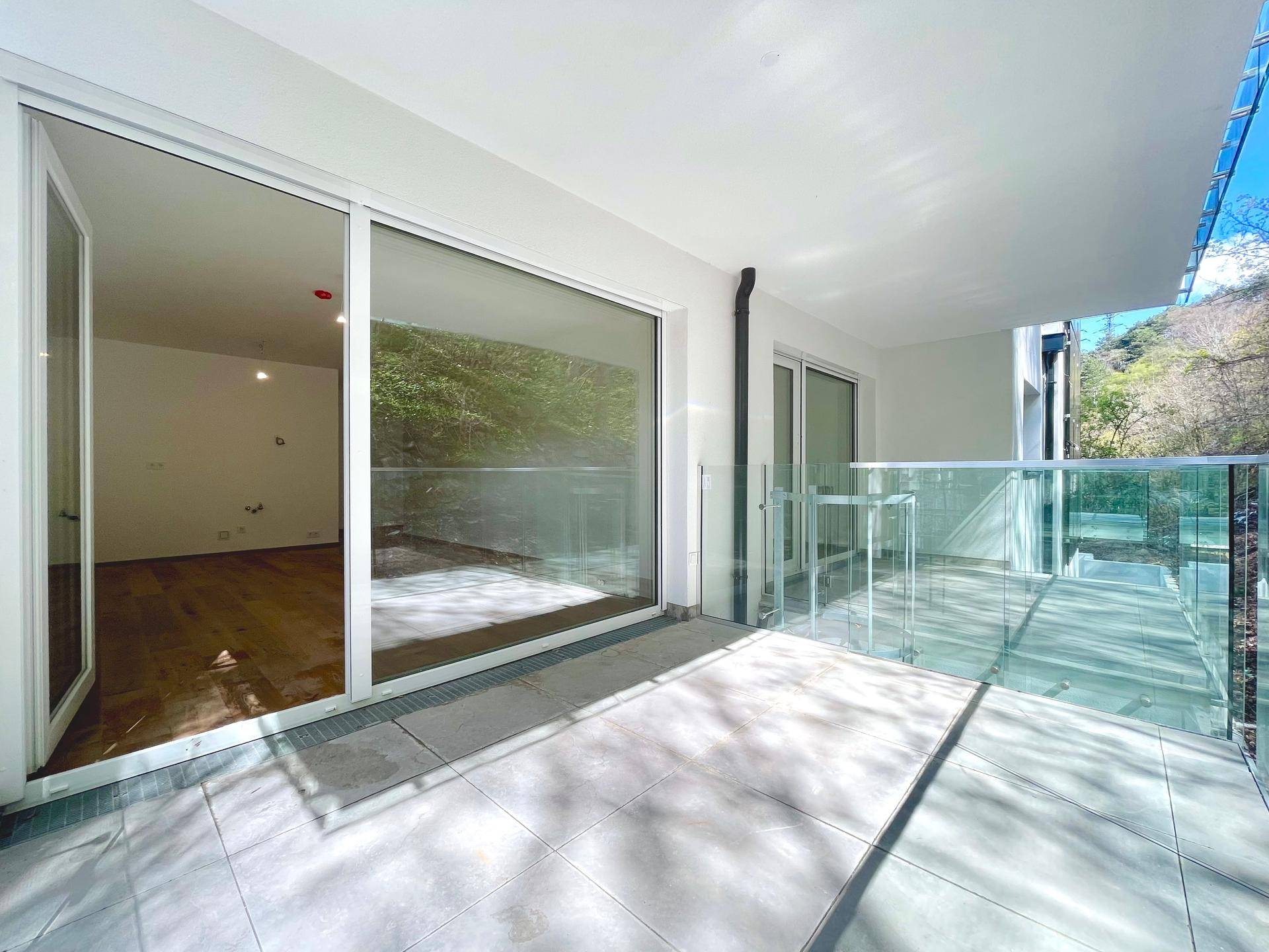 Schmaler Balkon mit Fliesenboden und Glasgeländer, bietet einen grünen Blick auf die umliegenden Bäume.