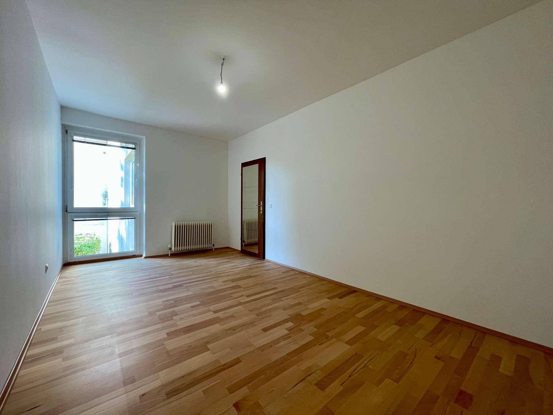 Lichtdurchflutetes Zimmer mit Holzboden und einem Fenster, das einen Blick ins Grüne bietet.