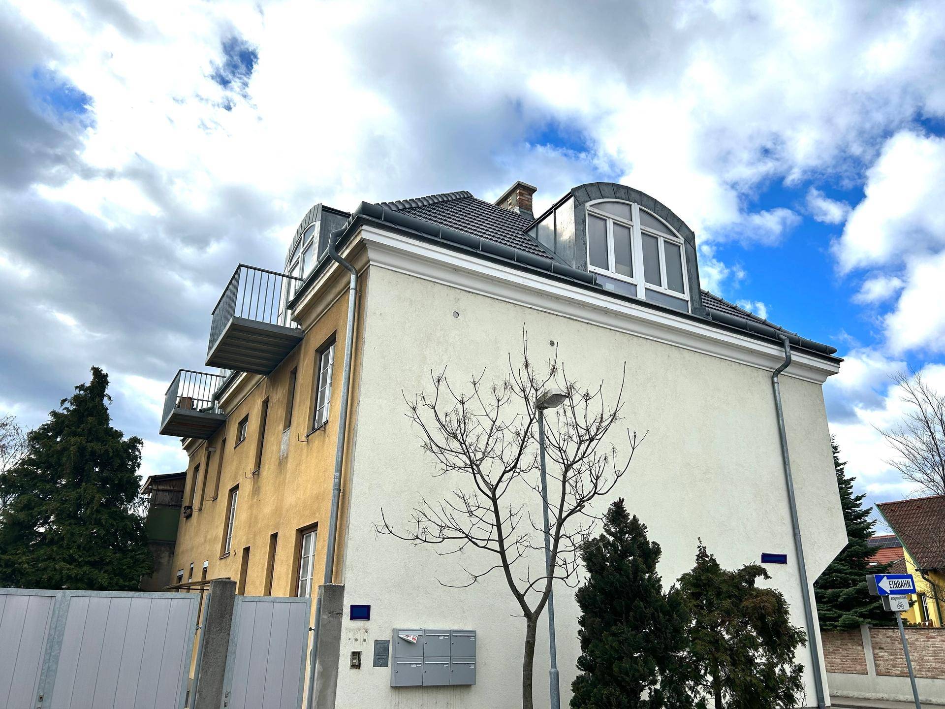 Seitenansicht des Gebäudes mit Mansarddach, Balkonen und einer hellen Fassade unter bewölktem Himmel.