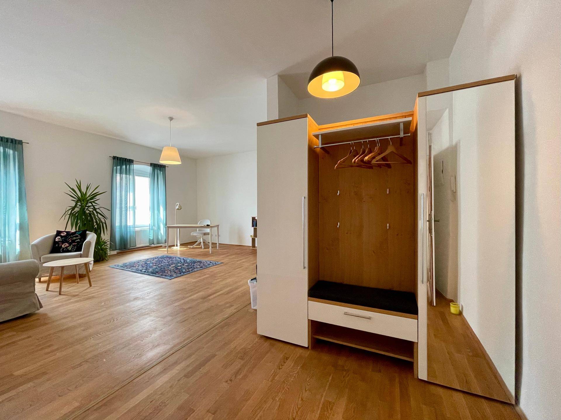 Geräumiger Wohnbereich mit Holzboden, Garderobe und einem Arbeitsbereich im Hintergrund.