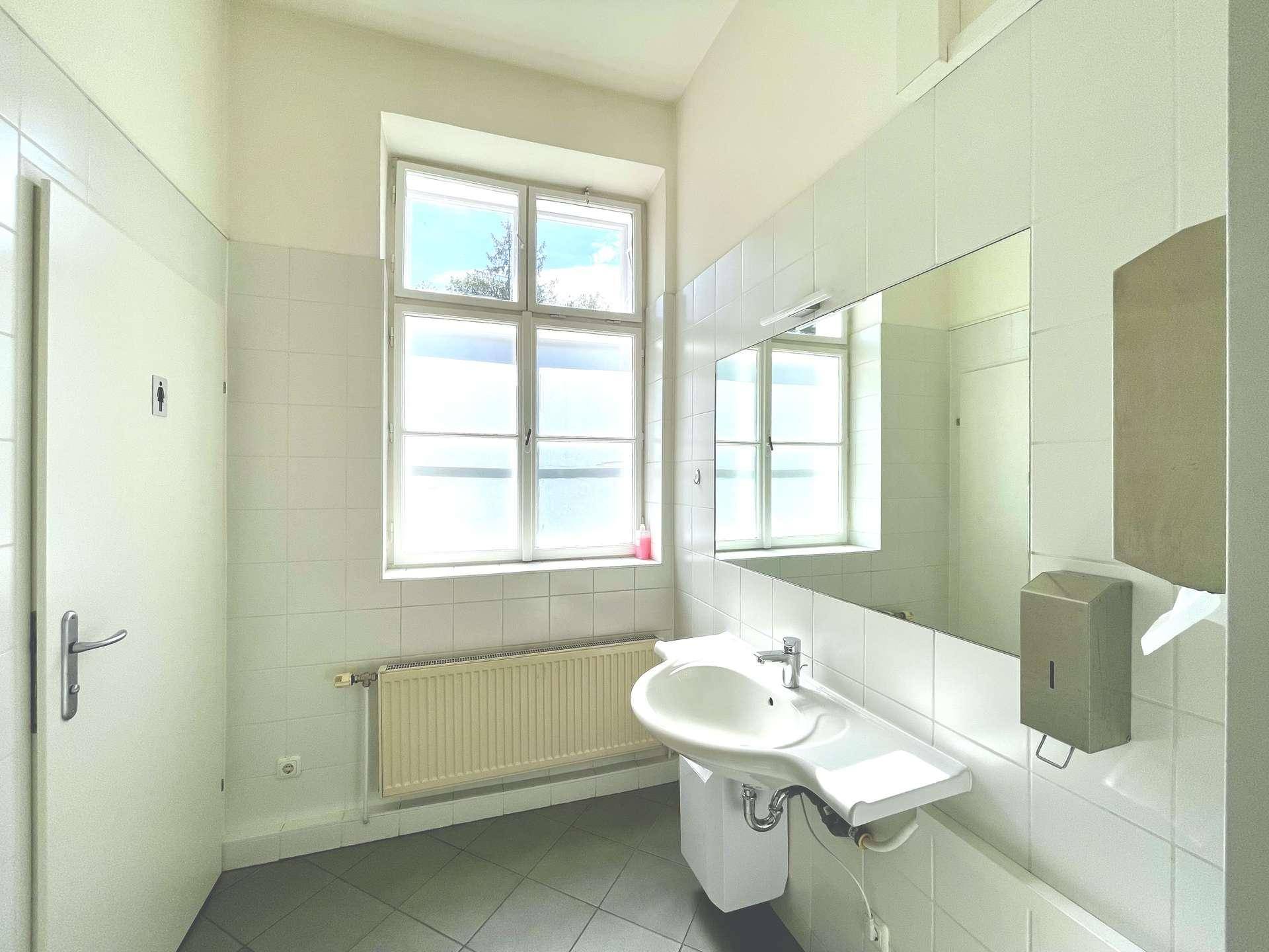 Helles Badezimmer mit weißen Fliesen, einem Fenster und einem Waschbecken mit Spiegel.