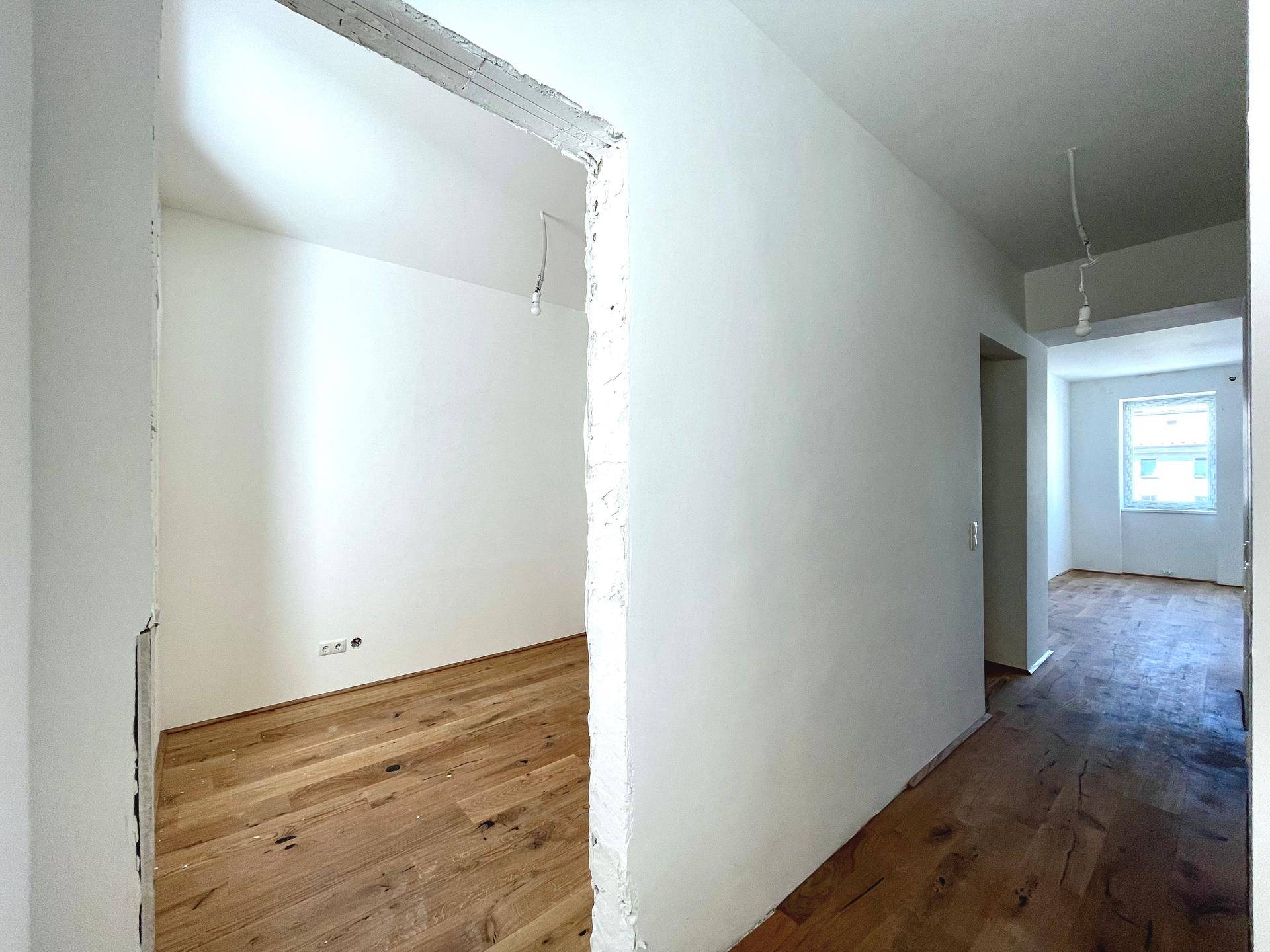 Heller Flurbereich mit Holzdielenboden, der zu einem weiteren unmöblierten Zimmer führt.