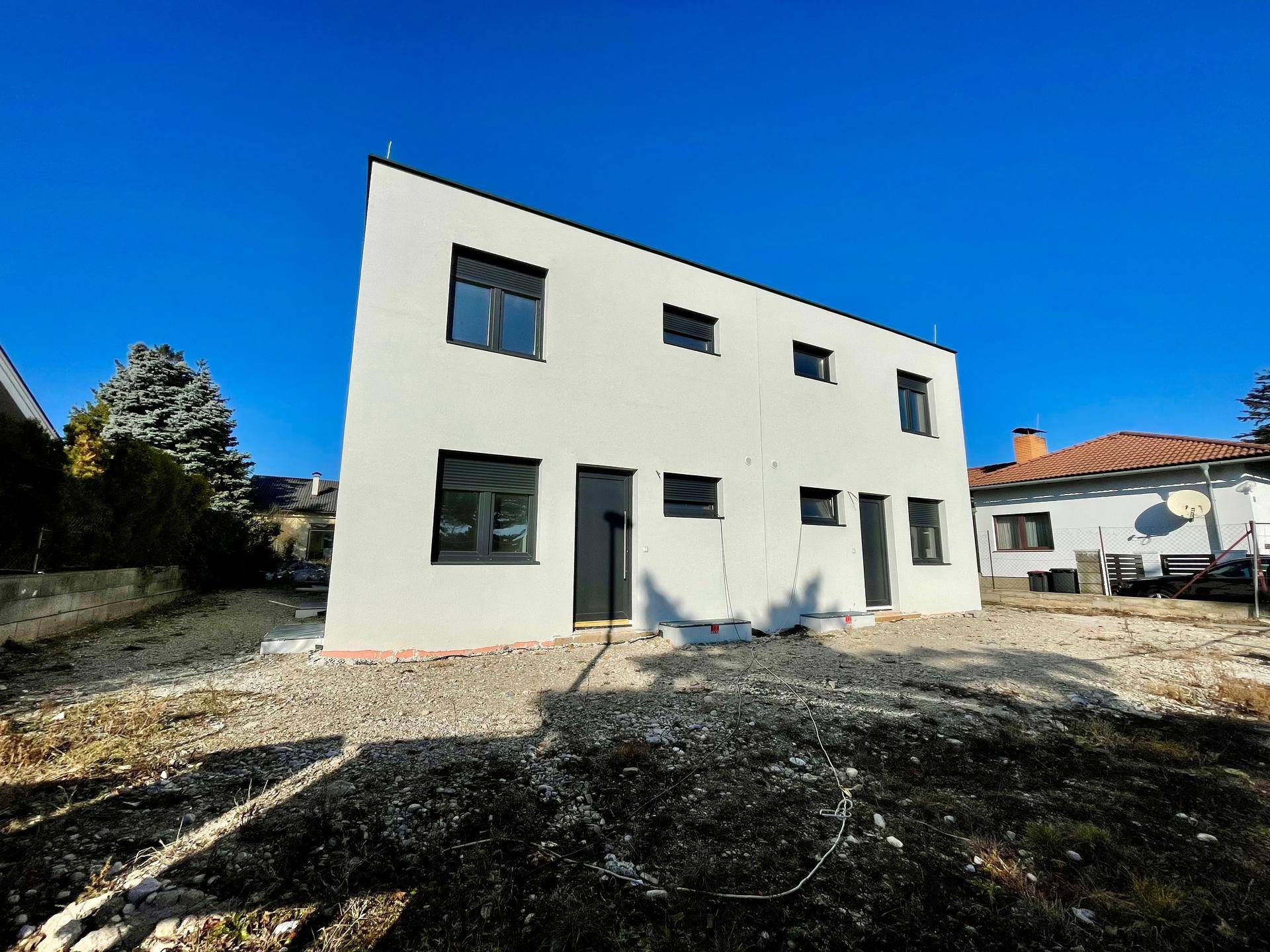 Modernes, weißes Neubauhaus mit dunklen Fenstern und Rollläden von der Seite.