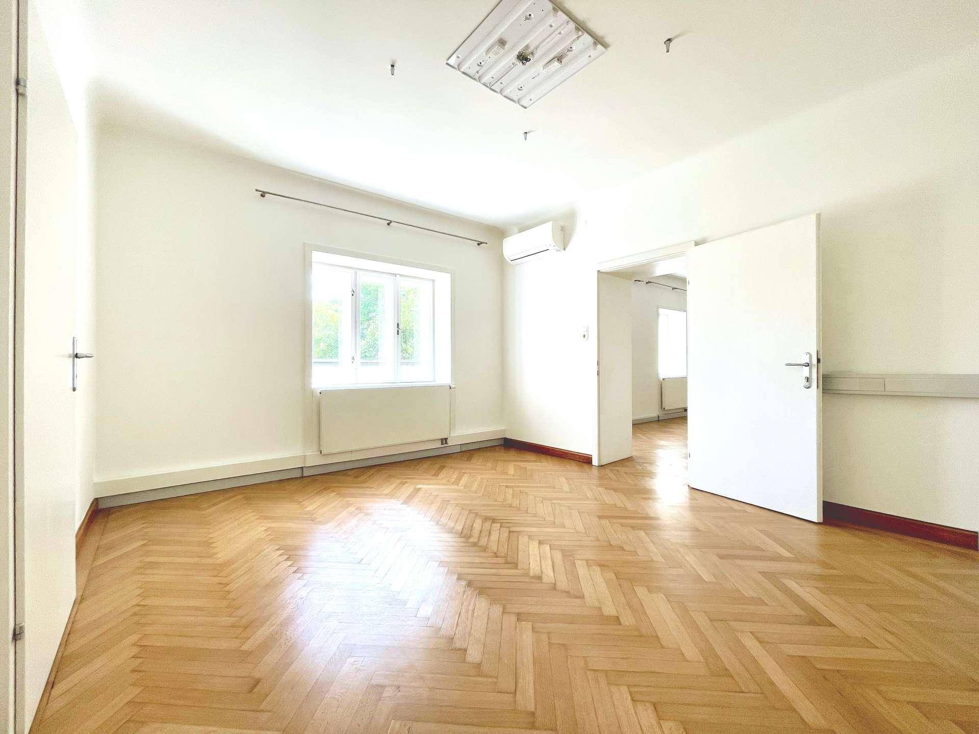 Geräumiger Raum mit Fischgrätparkett, einem großen Fenster und einer Klimaanlage.