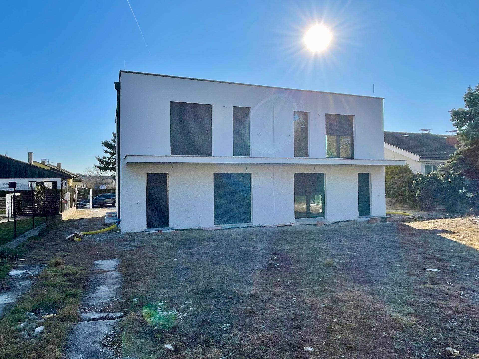 Modernes Einfamilienhaus mit weißer Fassade und dunklen Fensterrahmen unter strahlendem Sonnenschein.