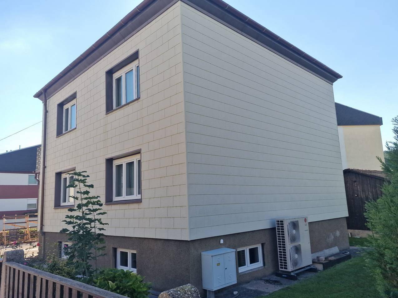 Modernes Gebäude mit heller Fassadenverkleidung und sichtbarer Wärmepumpe im Außenbereich.