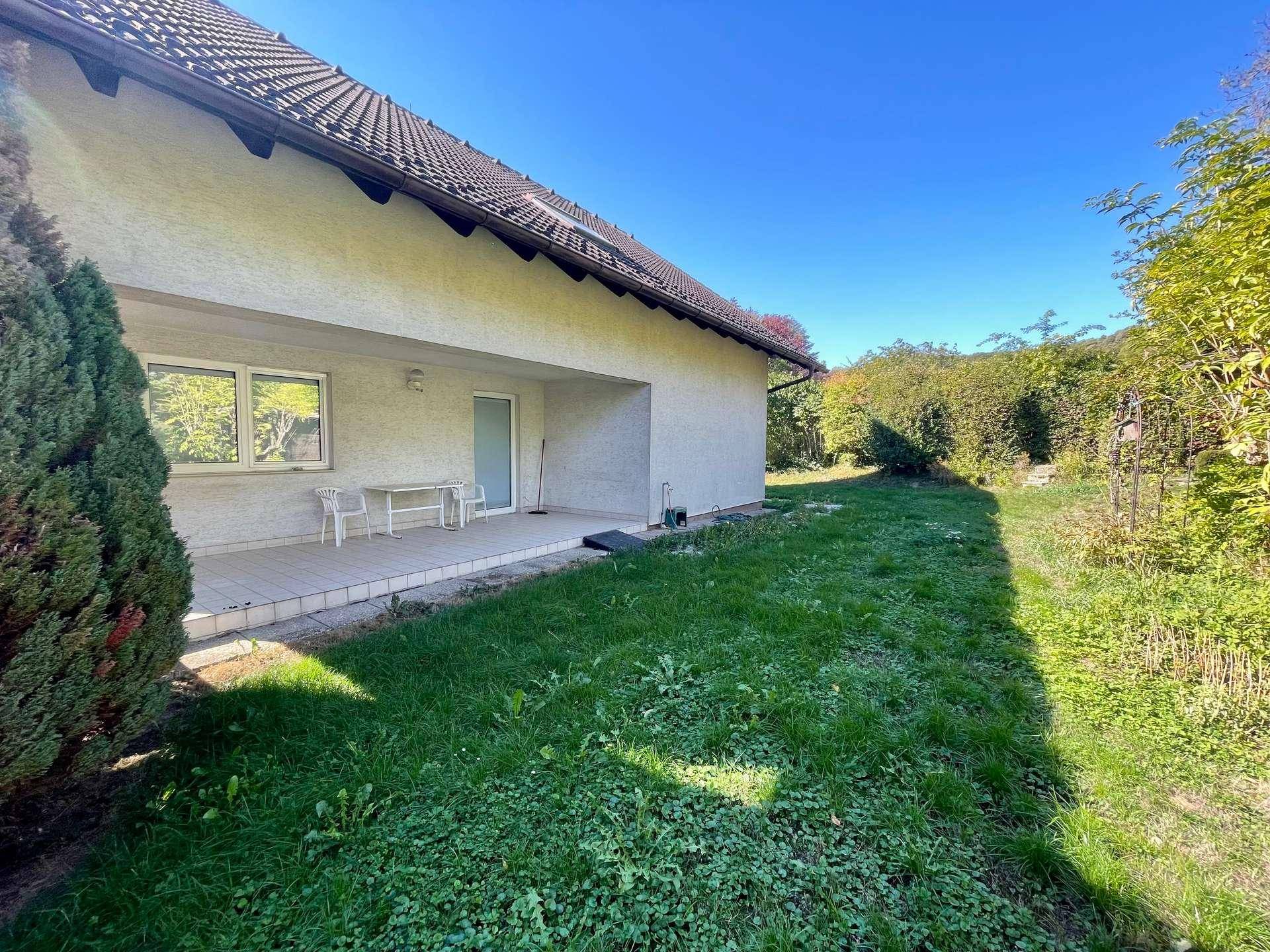 Überdachte Terrasse am Haus mit kleinem Tisch und Stühlen, umgeben von Grünflächen.