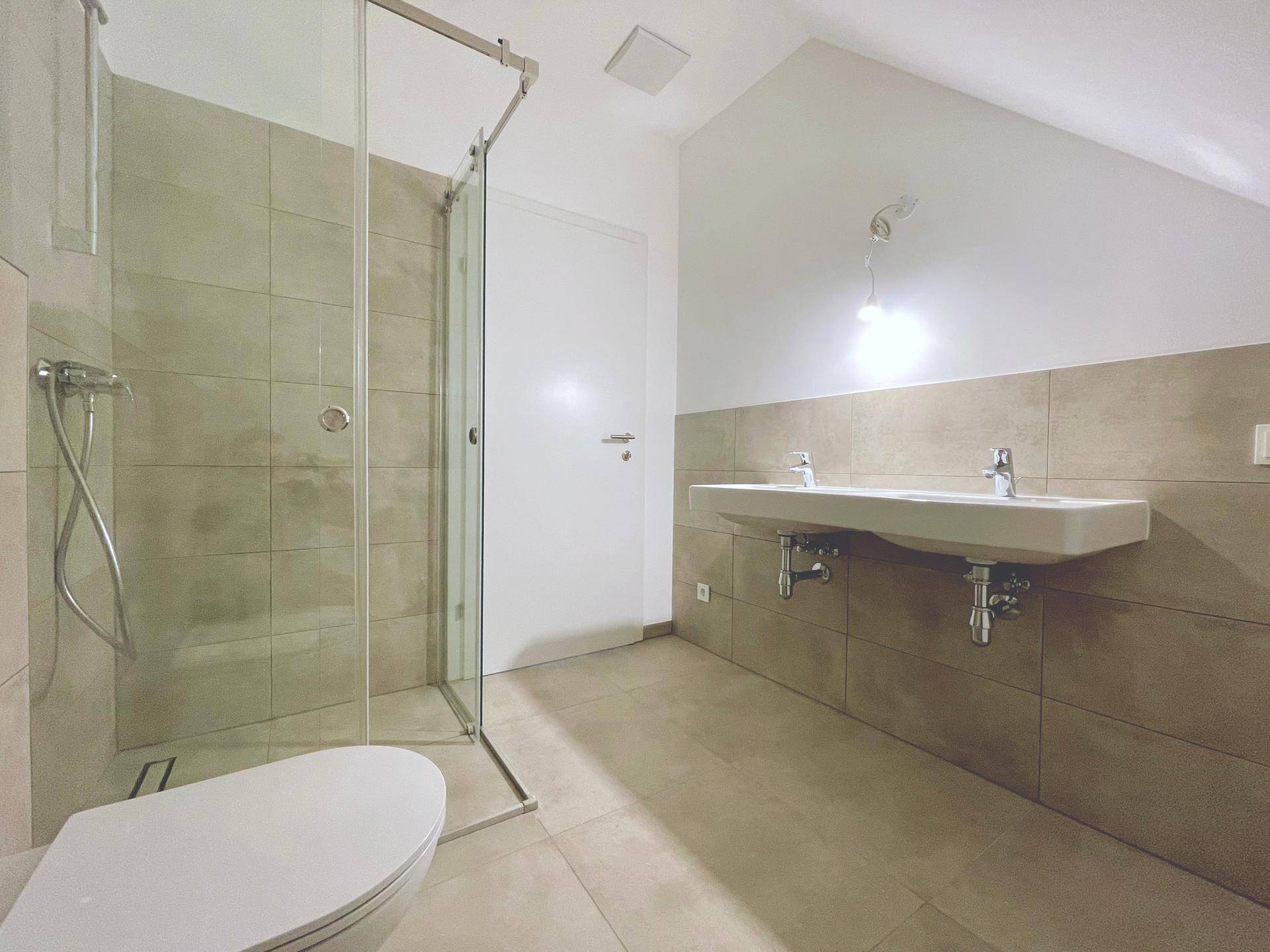 Modernes Badezimmer im Dachgeschoss mit Duschkabine, Wand-WC und Doppelwaschbecken, ausgestattet mit hellen Fliesen.