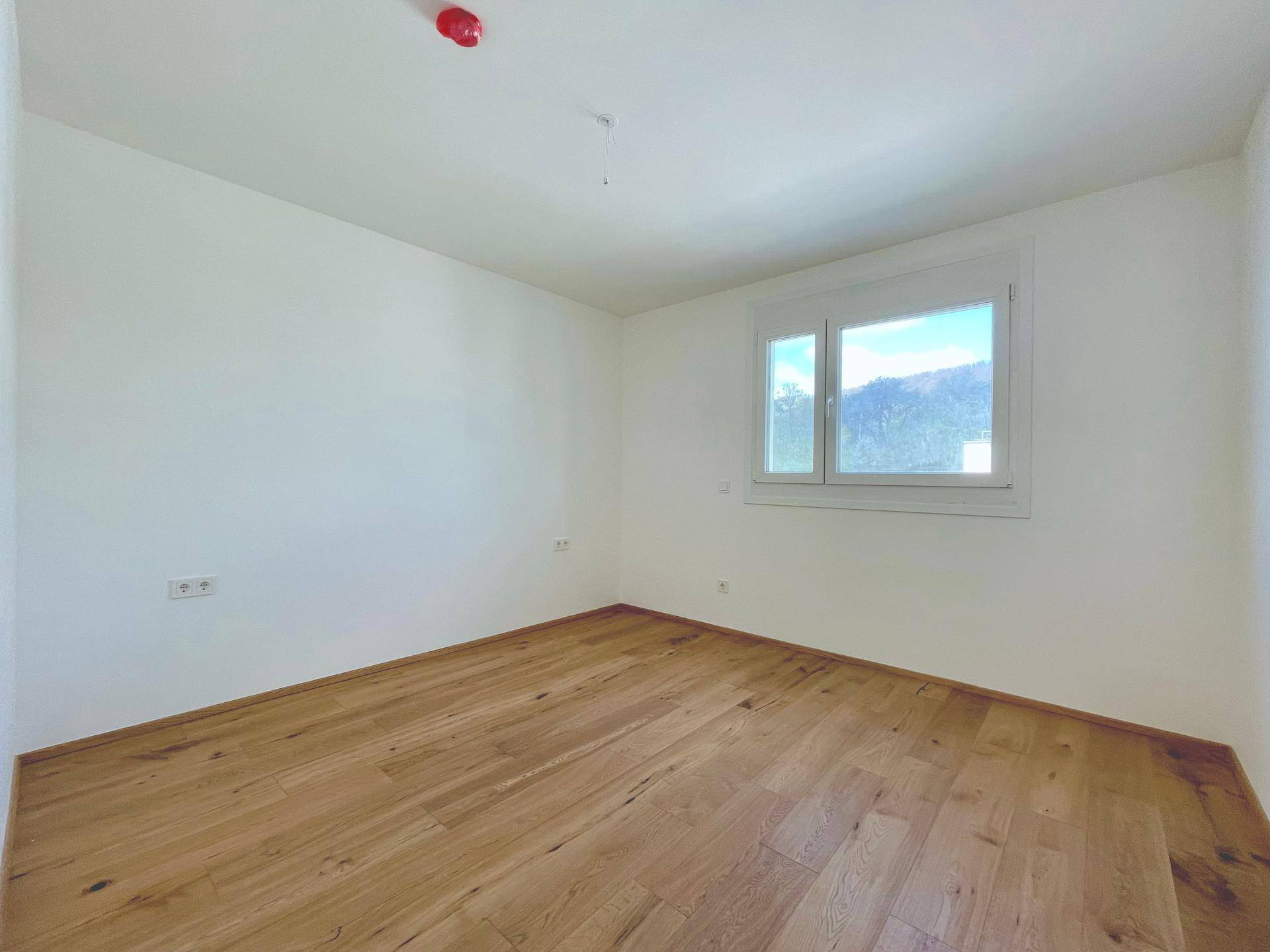 Schlafzimmer mit hellem Holzparkett und einem Fenster, das viel Tageslicht hereinlässt.