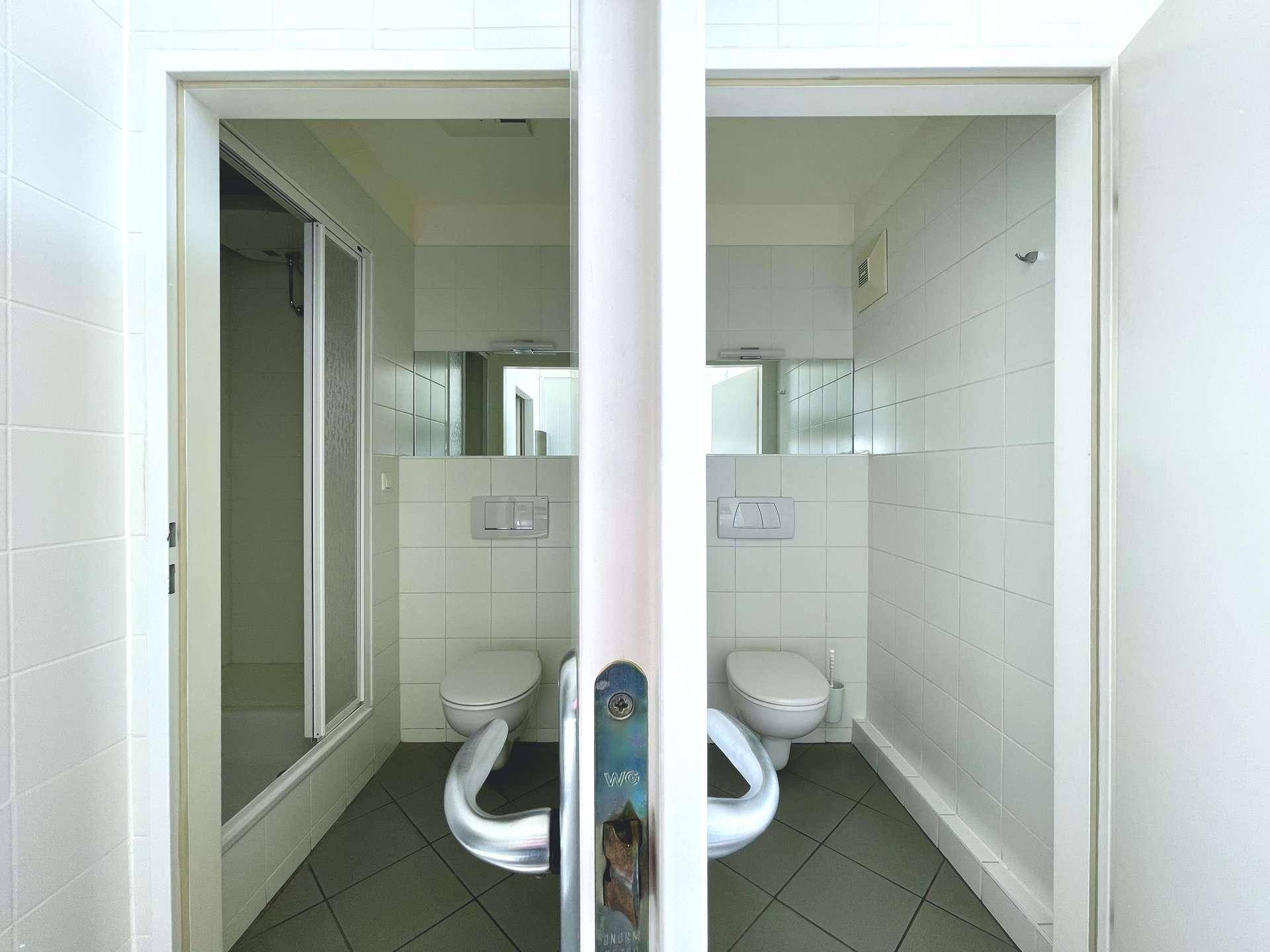 Zwei separate Toilettenbereiche mit weißen Fliesen, Dusche und Haltegriffen für Barrierefreiheit.