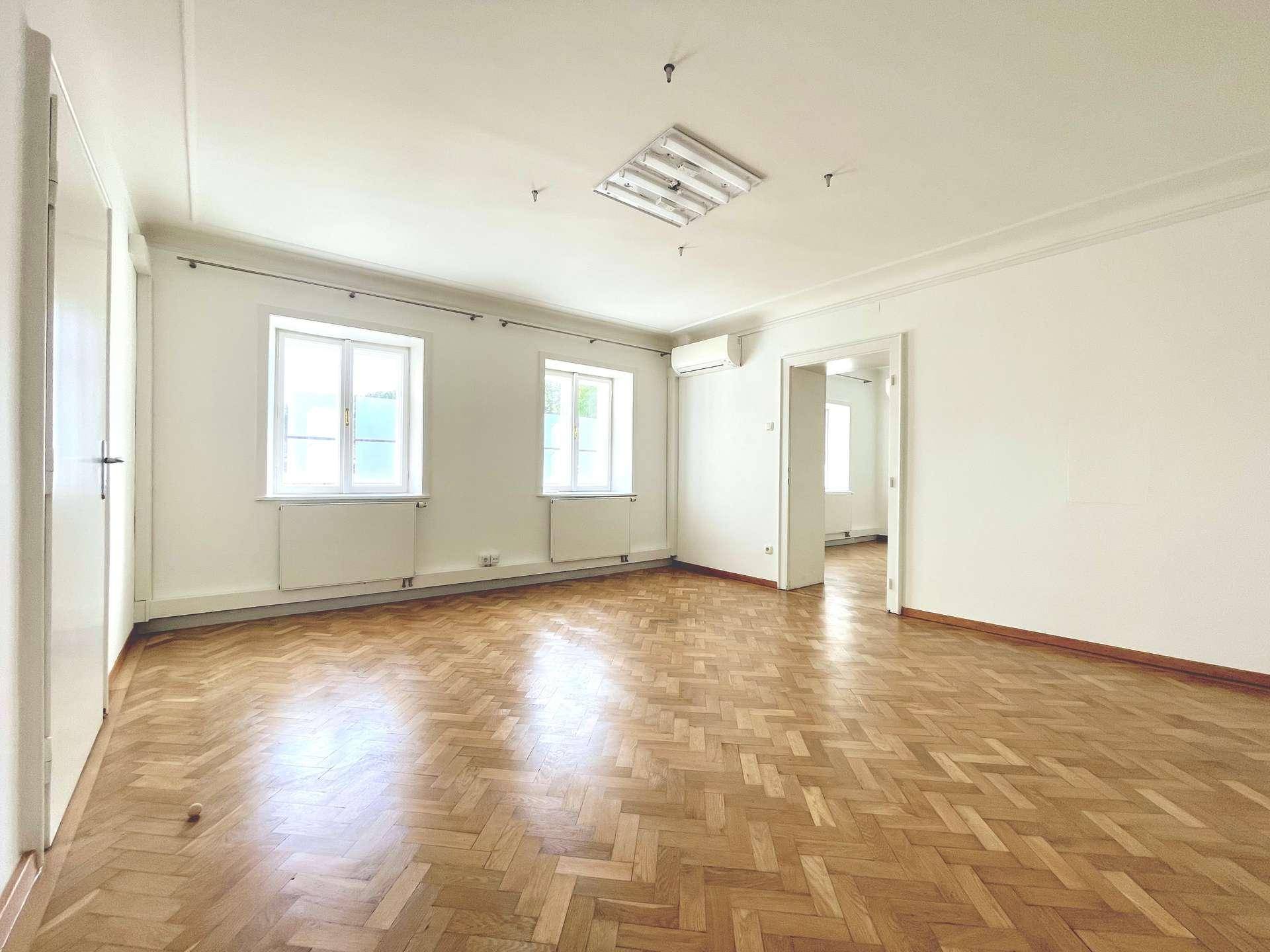 Heller Raum mit Fischgrätparkett, zwei Fenstern und einer Klimaanlage an der Wand.