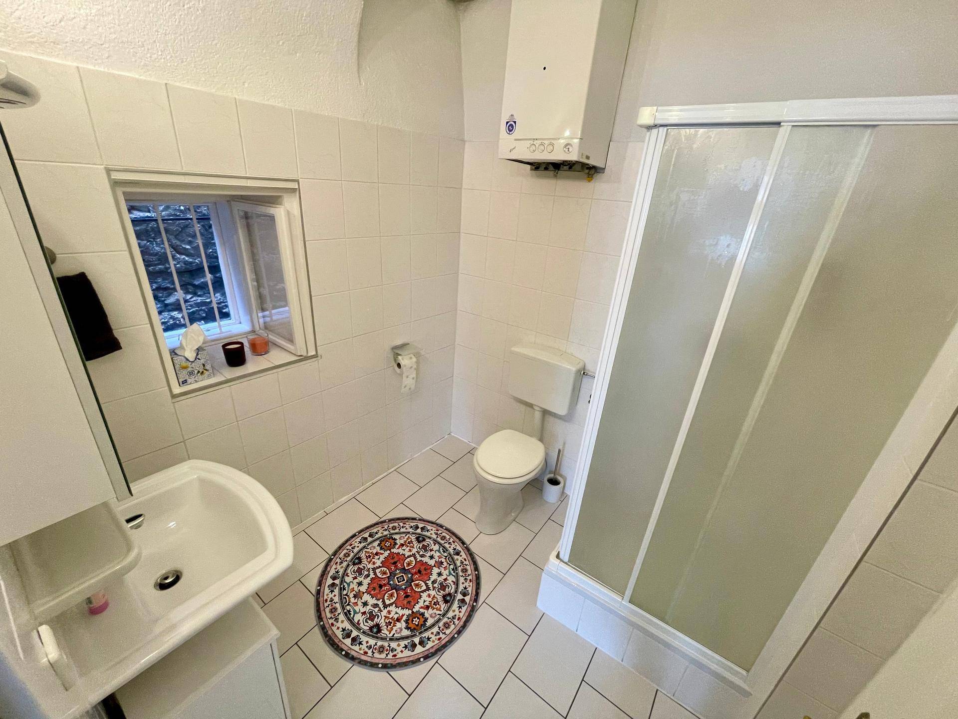 Gefliestes Badezimmer mit Dusche, Toilette, Waschbecken und einem kleinen Fenster.