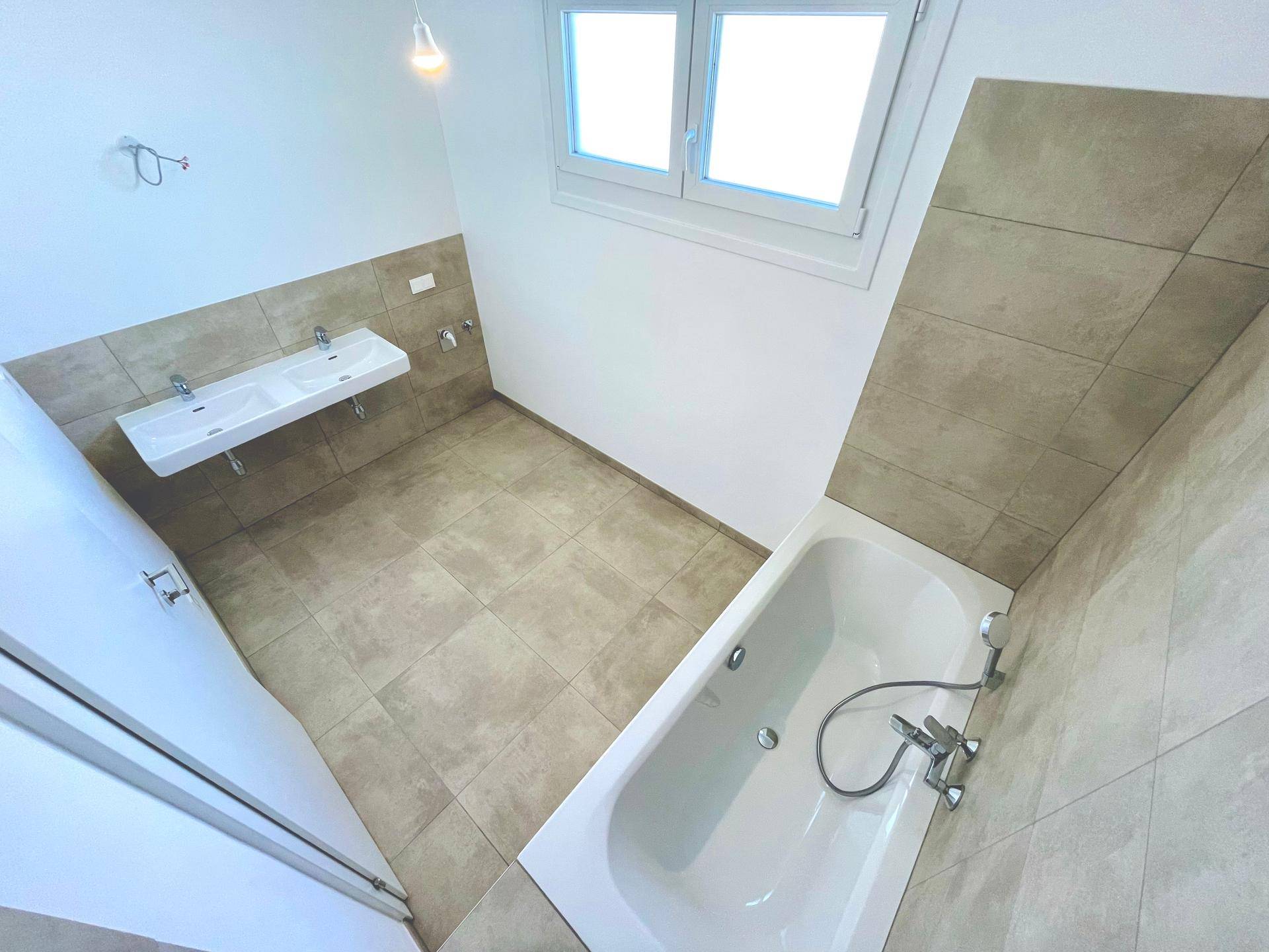 Modernes Badezimmer mit Badewanne, Doppelwaschbecken und Fenster, ausgestattet mit hellen Fliesen.