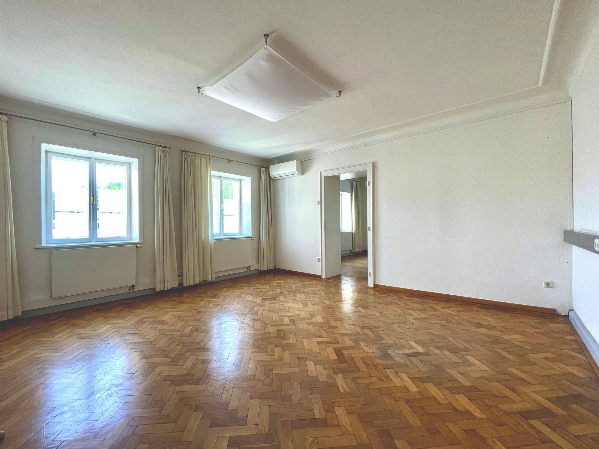 Heller Wohnraum mit Fischgrätparkett, hohen Decken, zwei großen Fenstern und Klimaanlage.