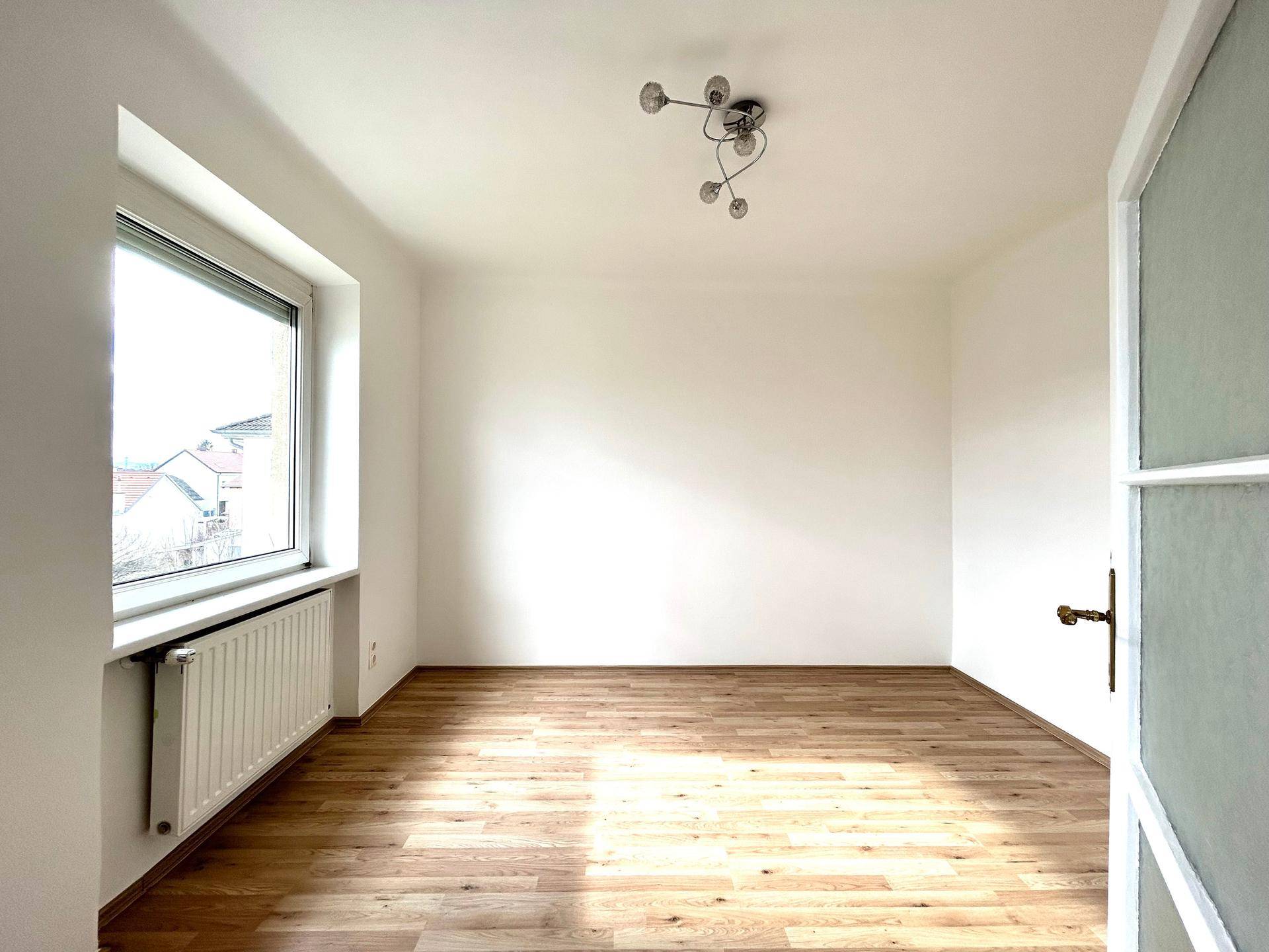 Helles Zimmer mit großem Fenster, Holzfußboden und moderner Deckenbeleuchtung.