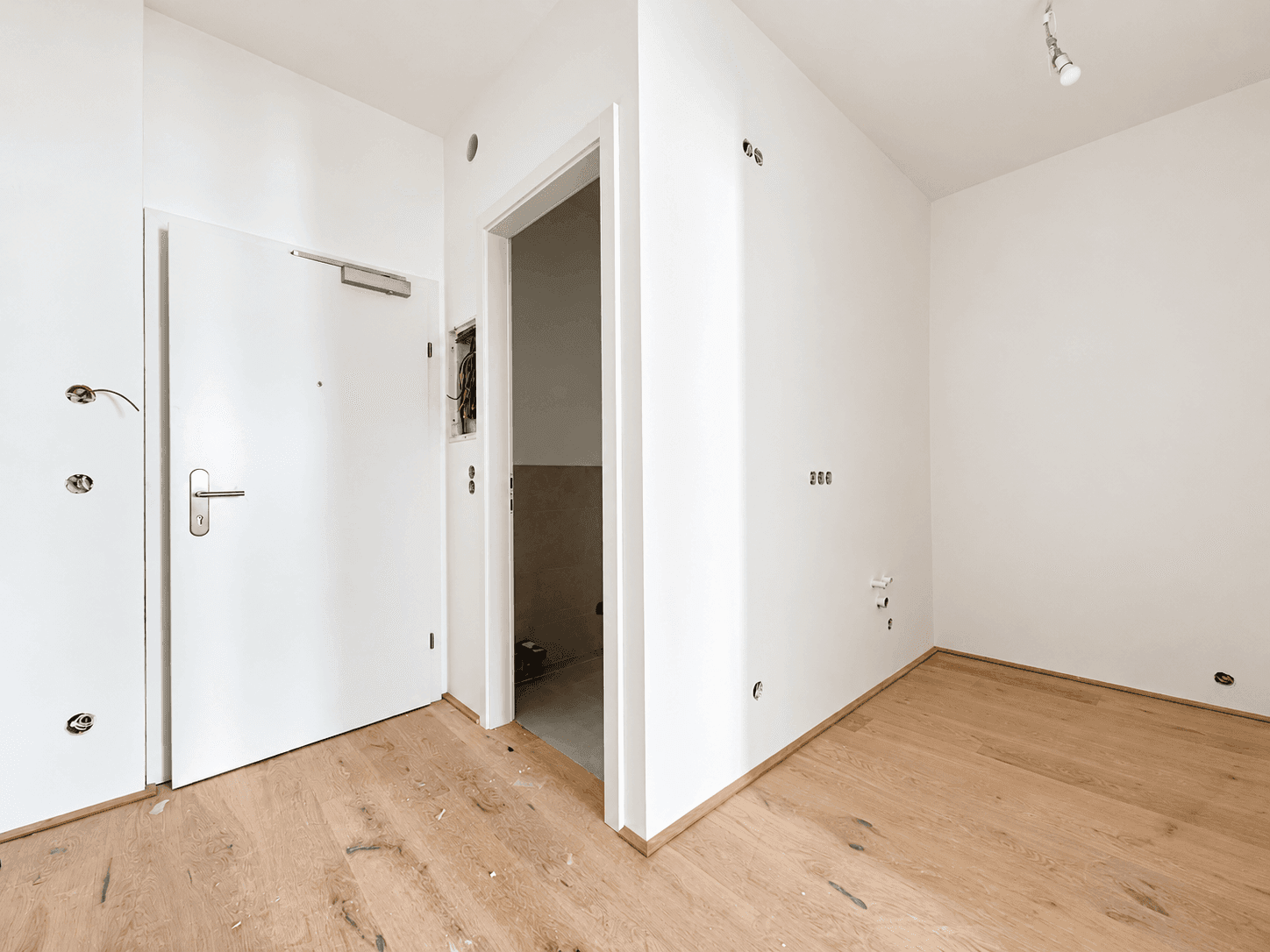 Eingangsbereich mit Holzboden und weißer Tür, bereit für die Installation von Beleuchtung und Schaltern.