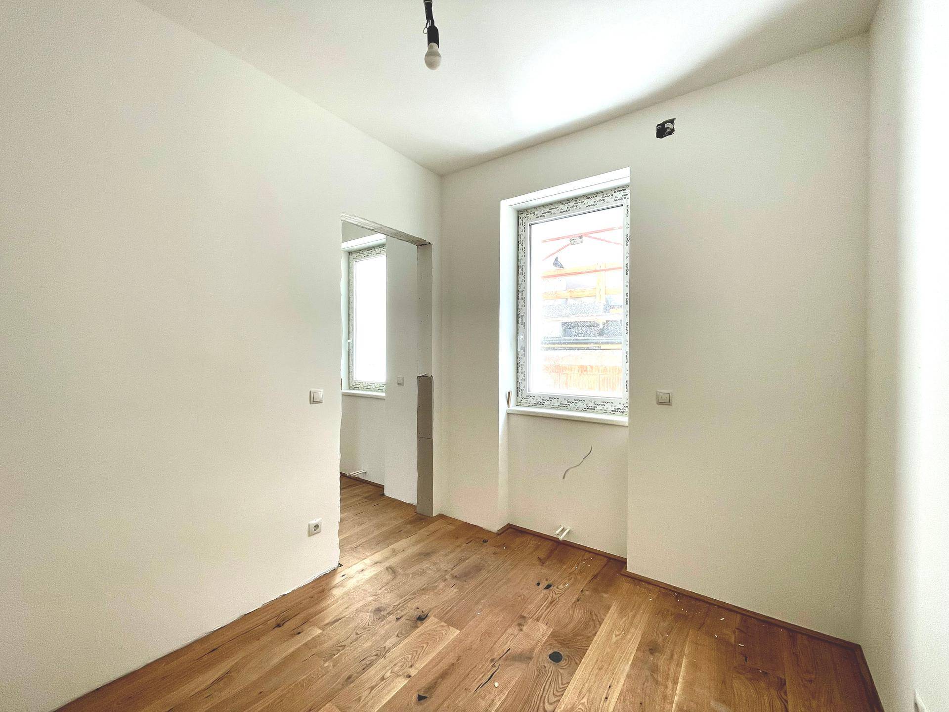 Helles Zimmer mit Holzboden und zwei Fenstern, die den Raum mit natürlichem Licht durchfluten.