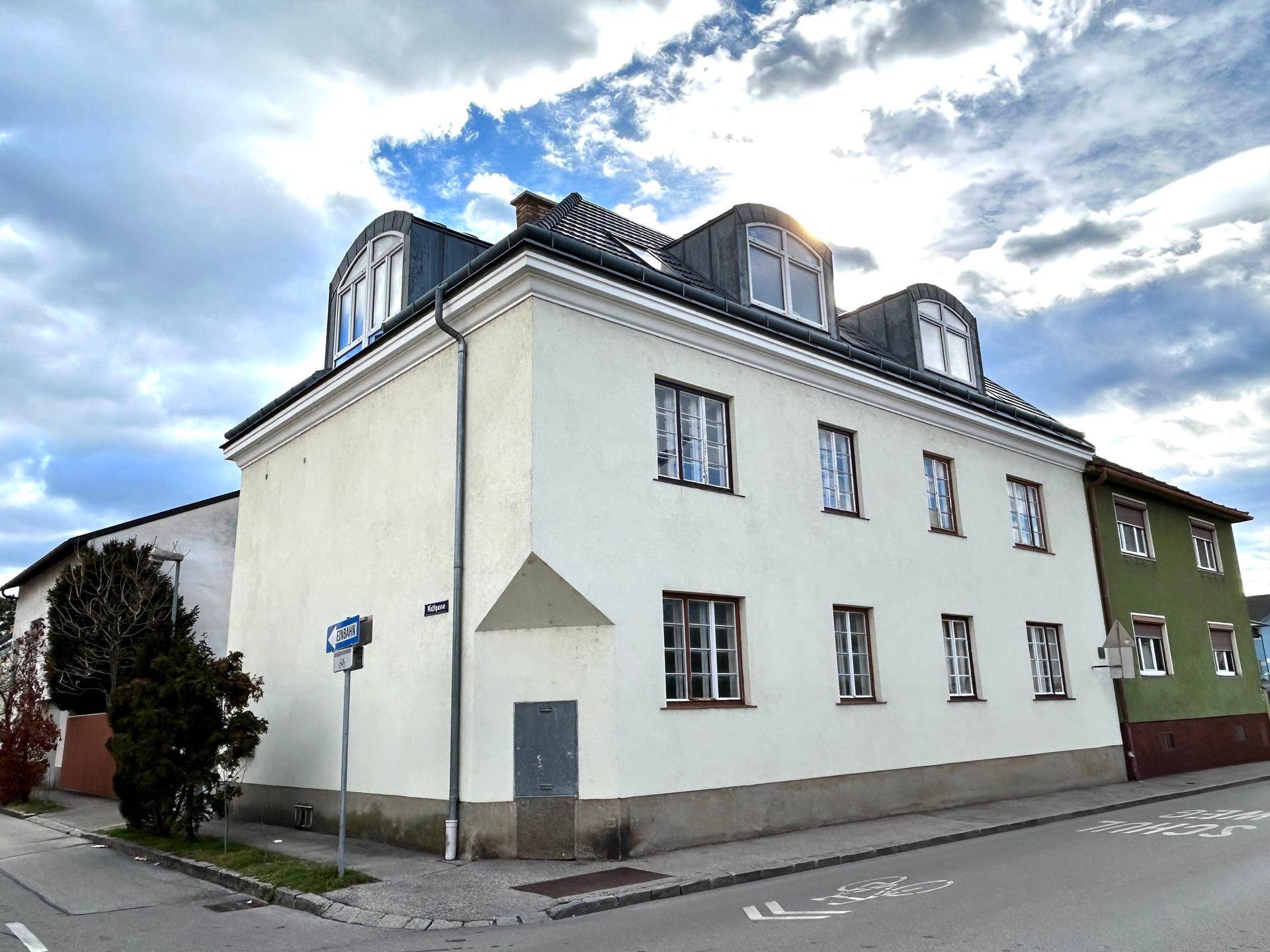 Außenansicht eines Gebäudes mit heller Fassade, Mansarddach und Gaubenfenstern an einer Straße.