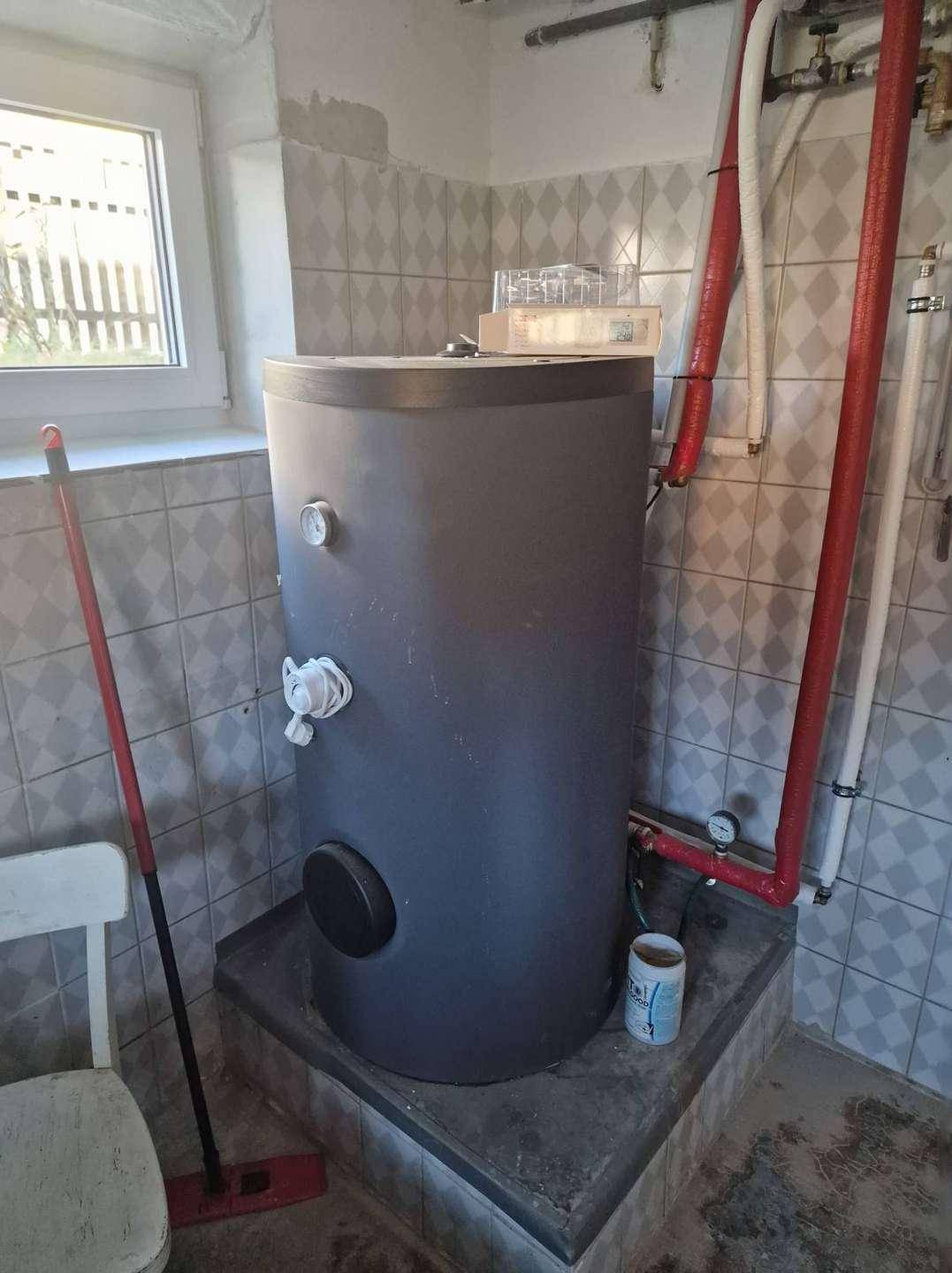 Großer Warmwasserspeicher in einem gefliesten Raum mit Fenster und roten Heizungsrohren.