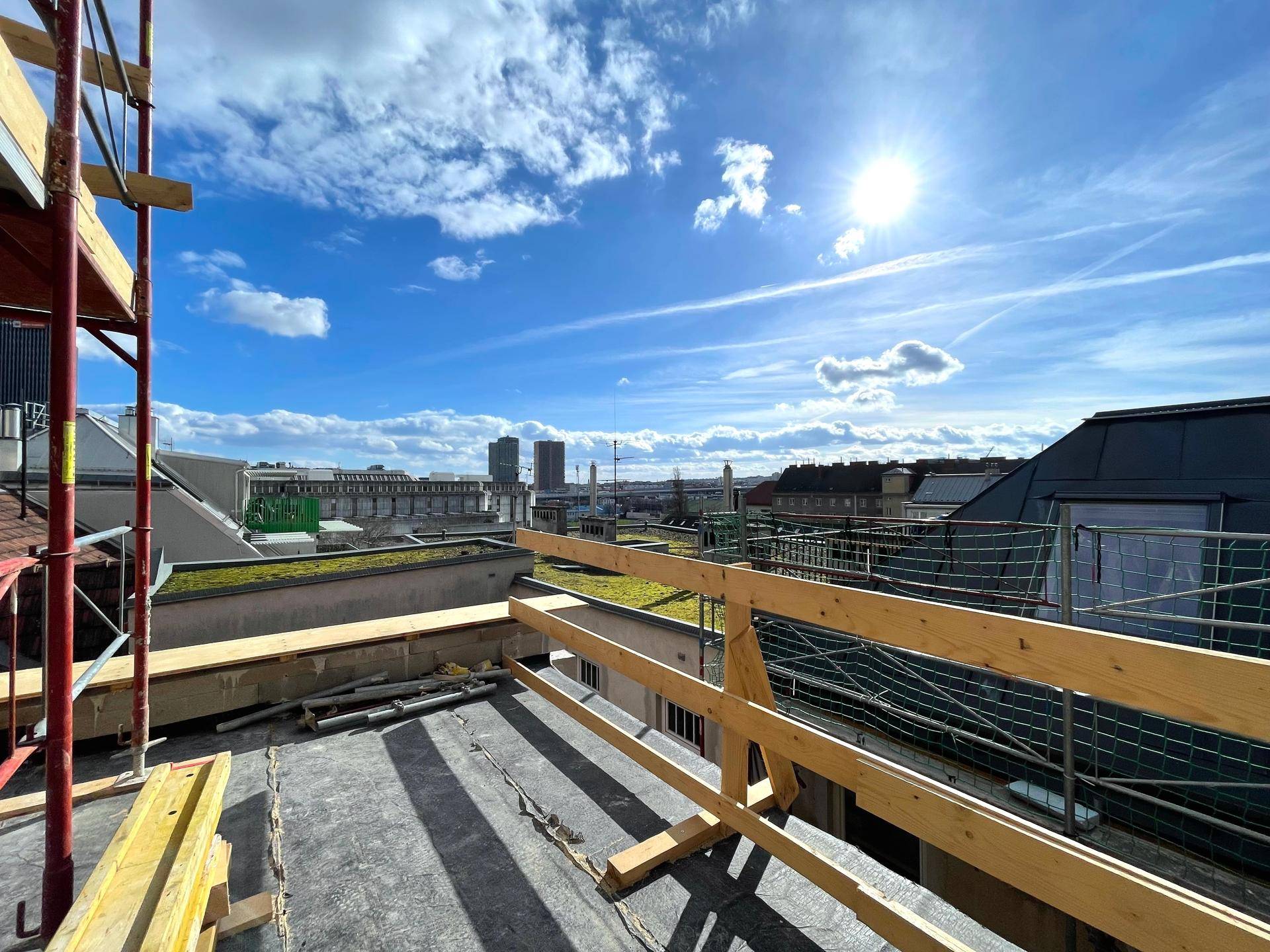 Dachterrasse im Bau mit Gerüst und weitem Blick über die Stadt unter strahlendem Himmel.