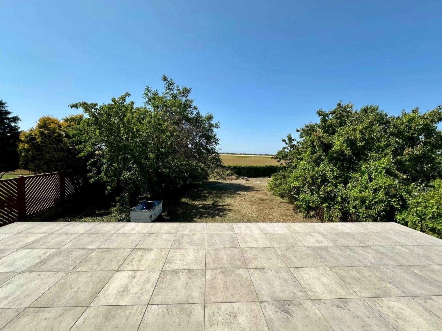 Weitläufiger Blick von der Terrasse über den Garten auf Felder und Bäume unter blauem Himmel.